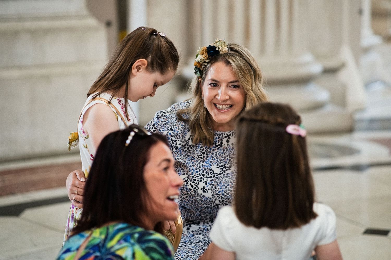 Women and girls in floral outfits share a joyful moment together in an elegant columned hall.