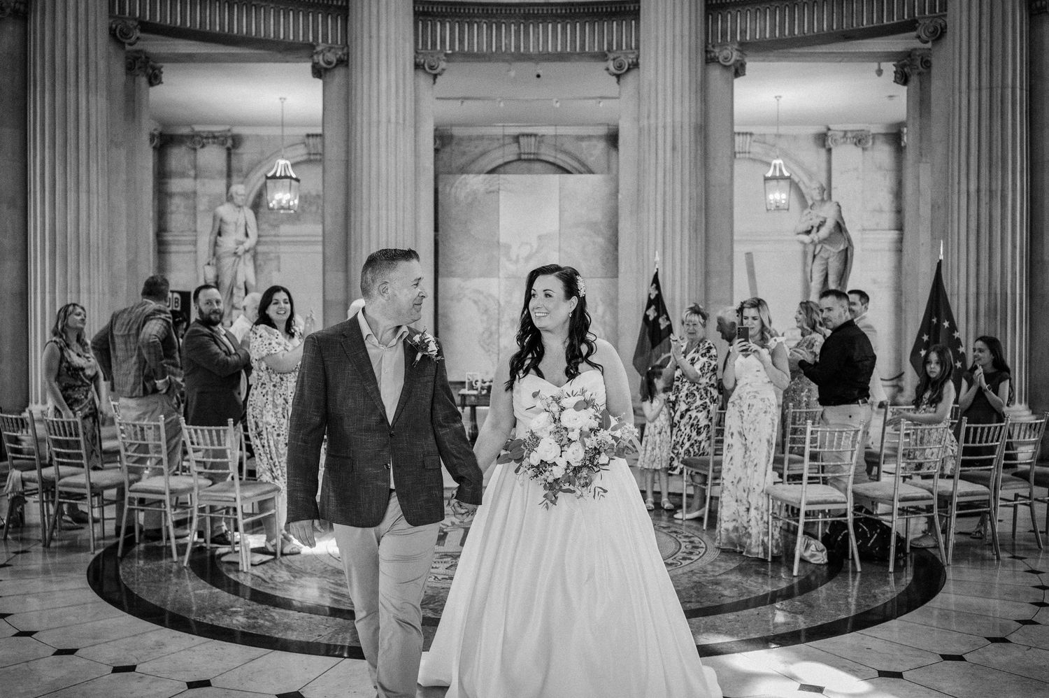 Black and white photo of a bride and groom walking hand in hand after their wedding ceremony in an elegant columned hall.