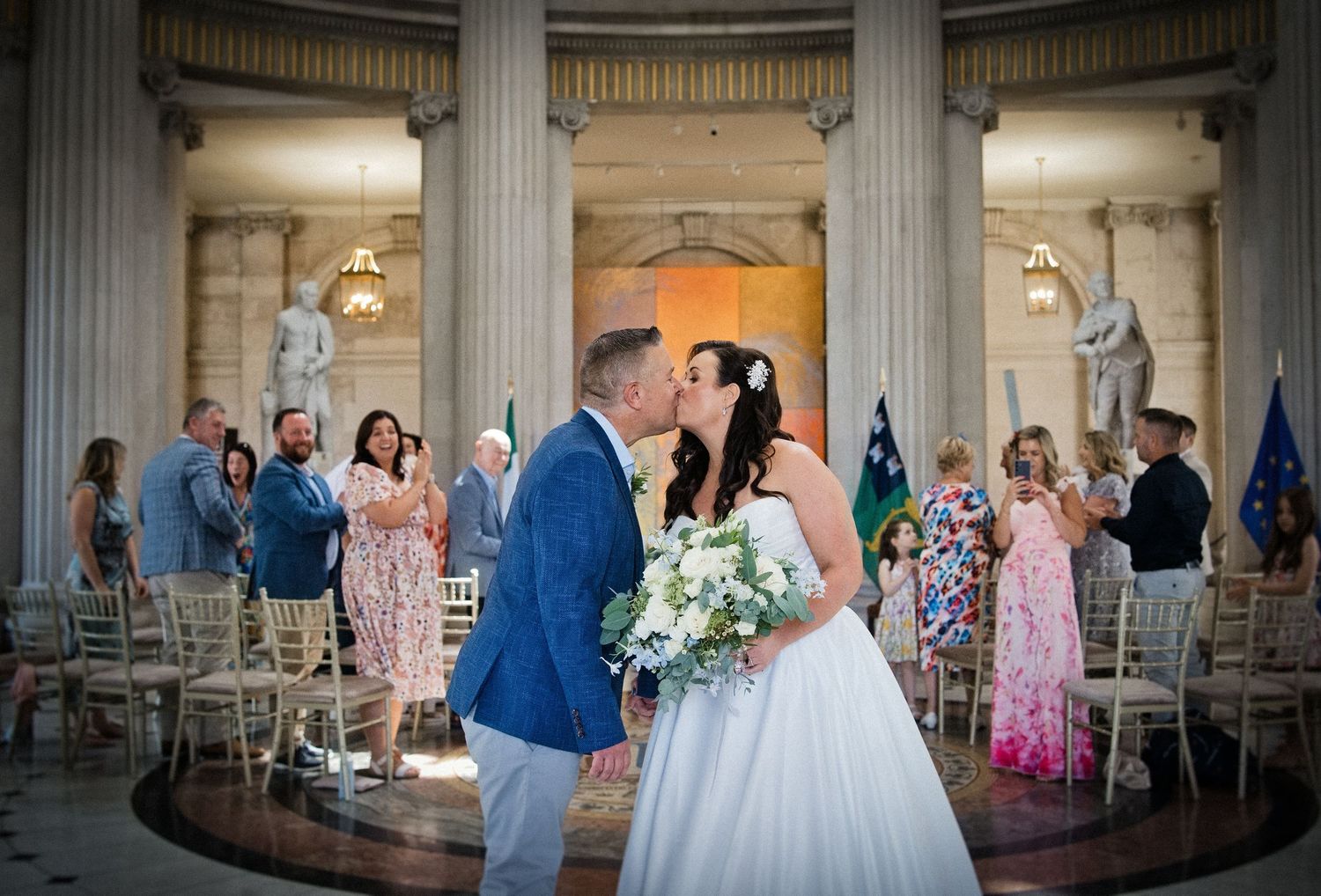 Bride and groom share a first kiss at elegant wedding ceremony in grand columned hall with guests applauding.