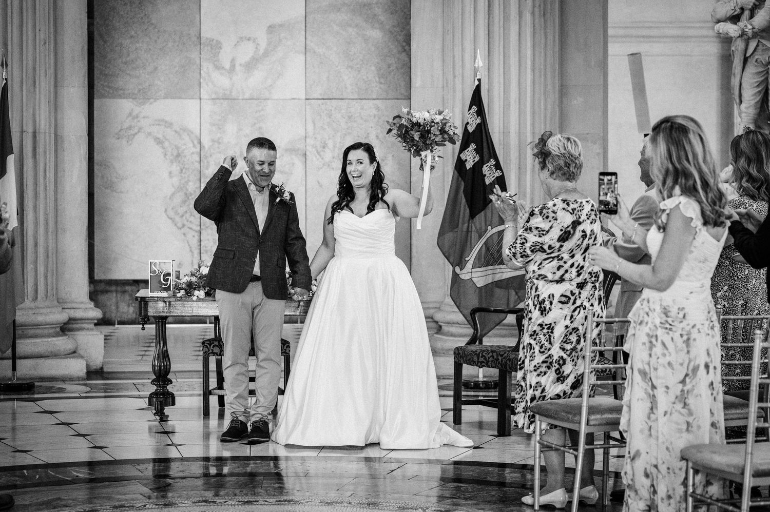 Bride and groom celebrate at wedding ceremony in elegant columned venue, guests capturing the joyful moment.