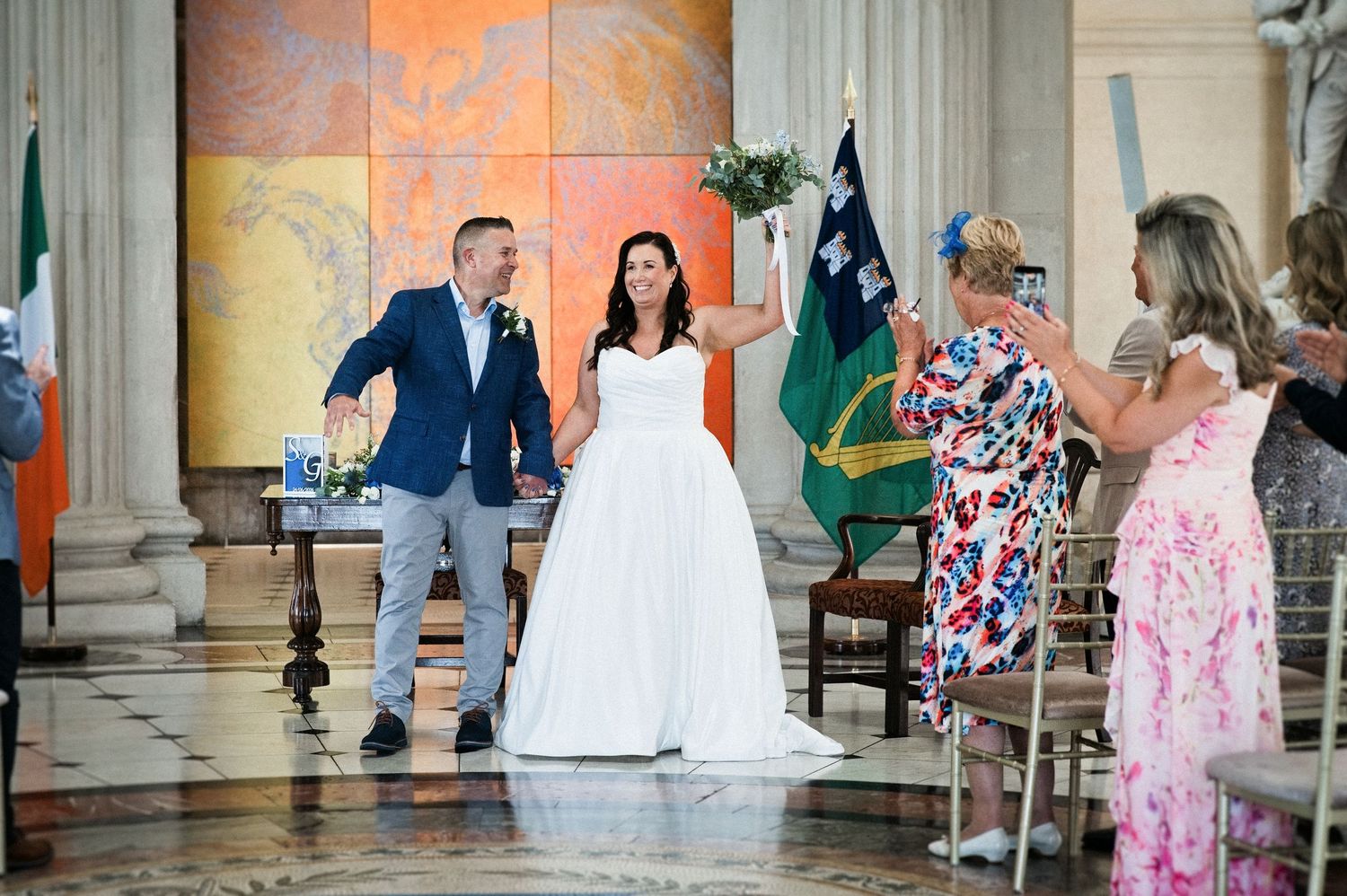 Bride and groom celebrate their wedding ceremony in an elegant hall with guests clapping and Irish flag in background.
