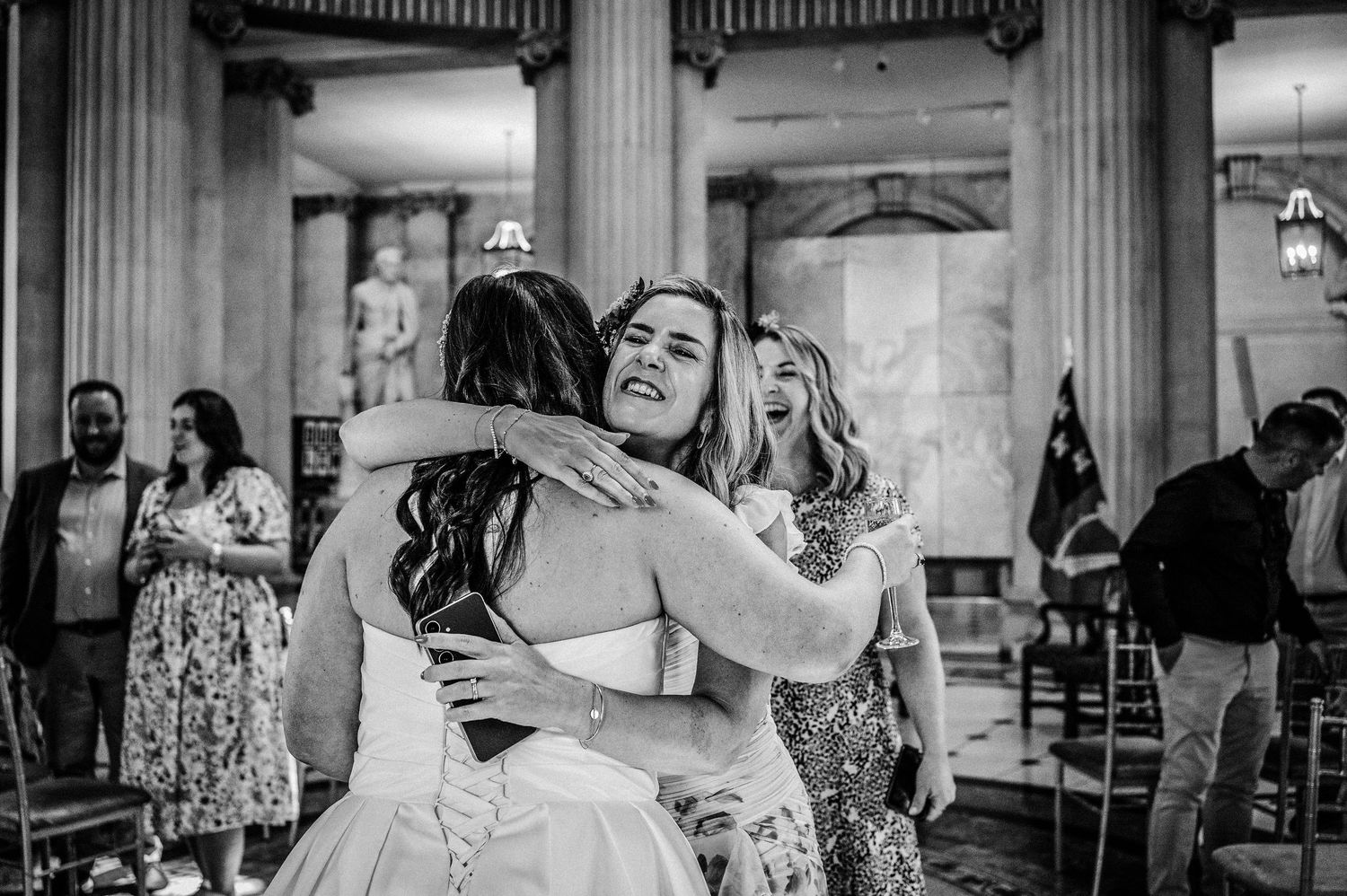 Black and white photo of bride hugging guests at wedding reception inside elegant columned venue.