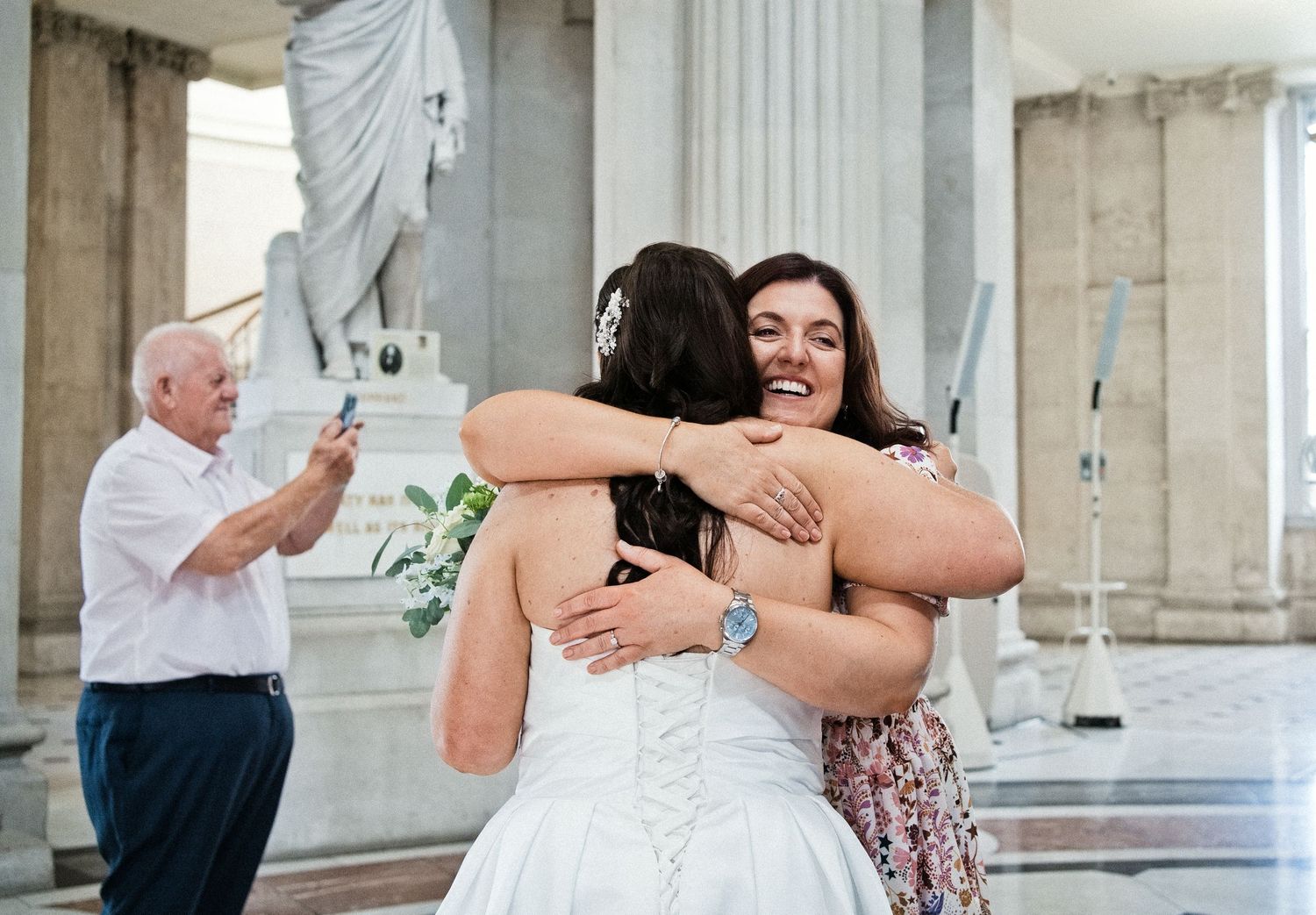 Bride and woman share warm hug at wedding venue with classical columns while man photographs in background.