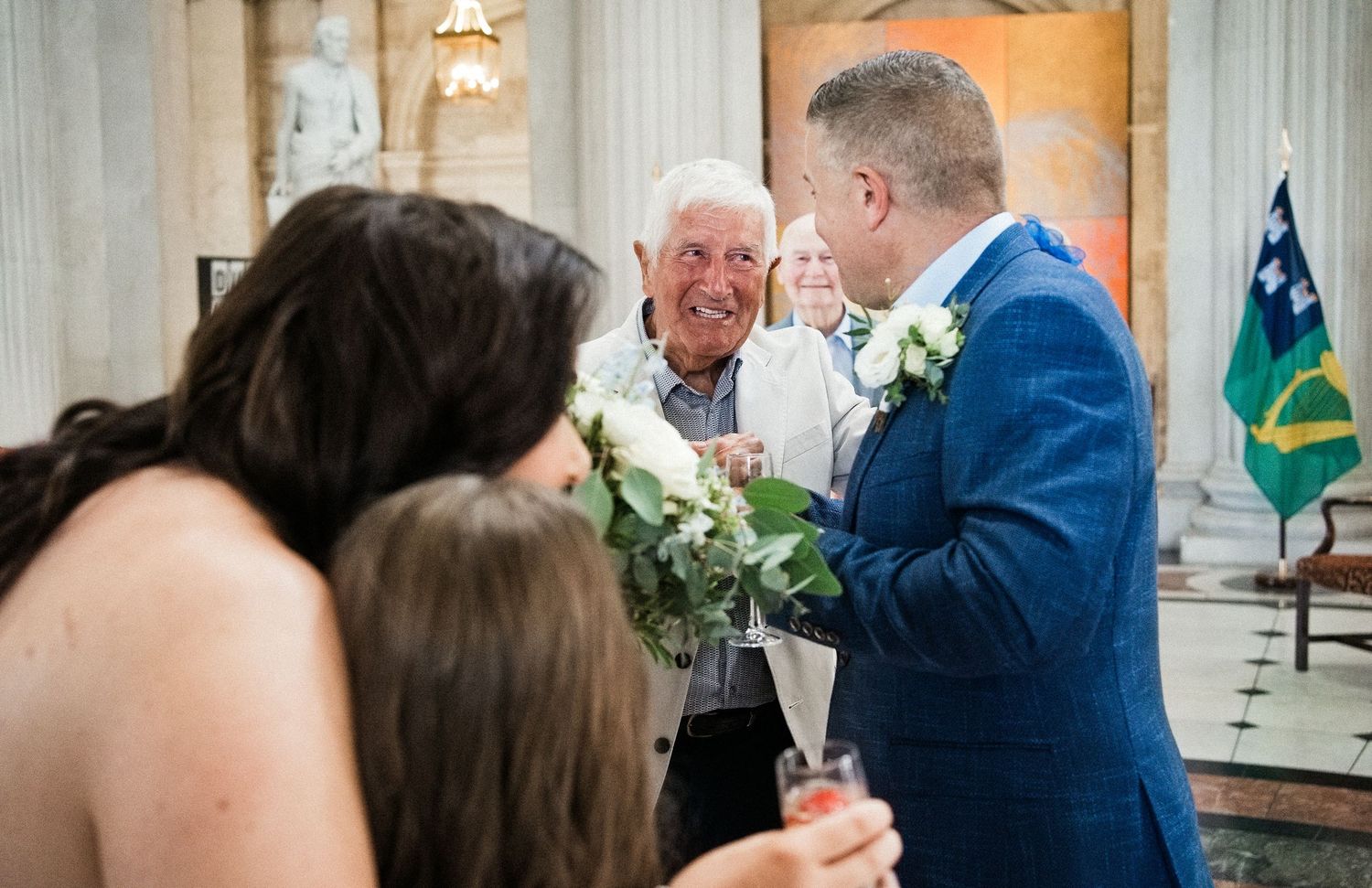 Elderly man smiling and greeting wedding couple in blue suit with white boutonniere inside elegant venue.