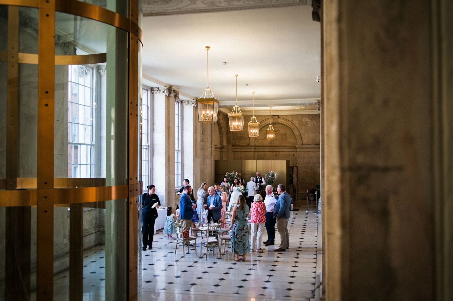 Visitors gather in an elegant grand hall with marble floors, tall columns, and ornate chandeliers in a historic building.