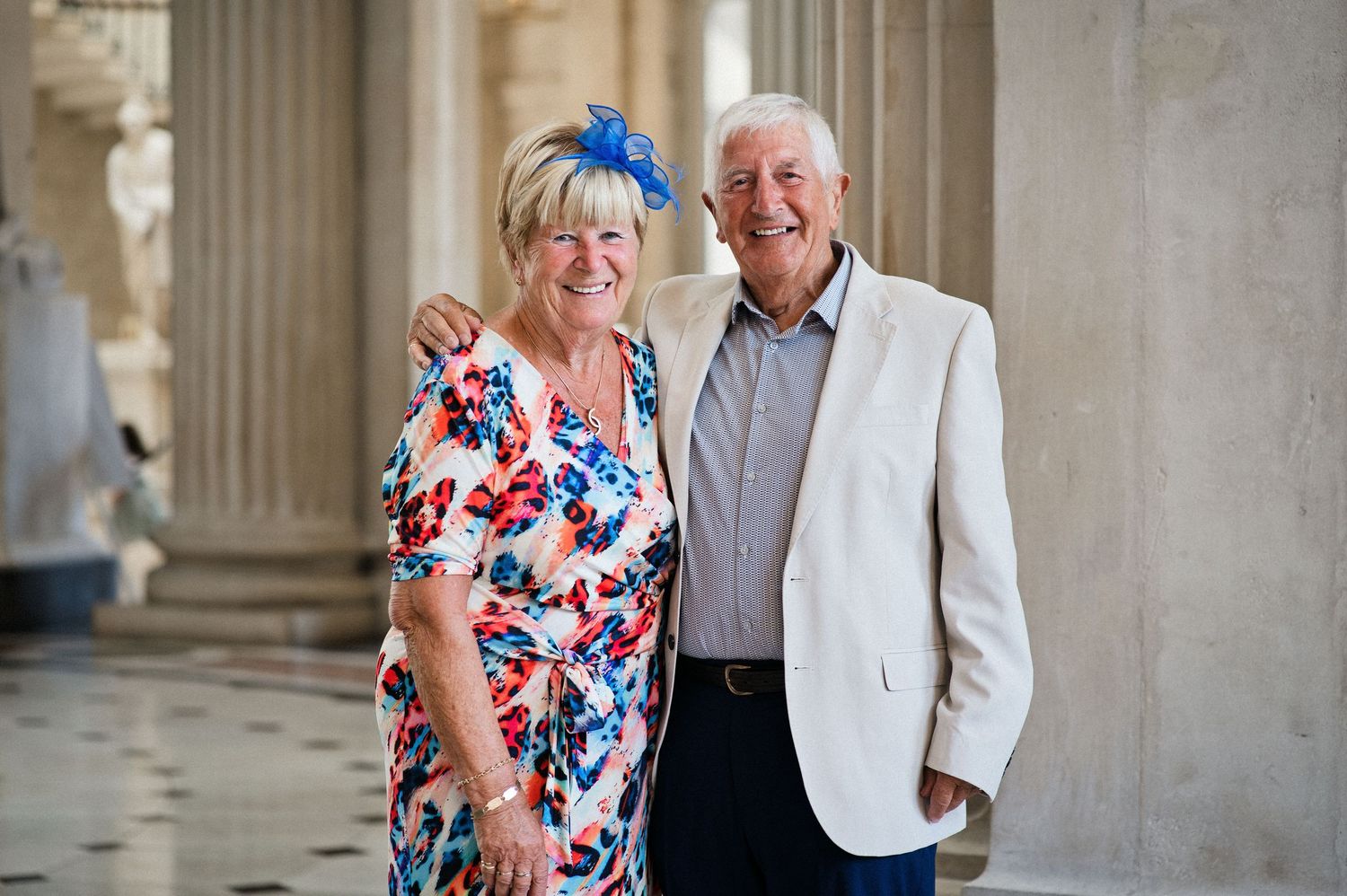 Smiling elderly couple posing together in elegant columned hall, woman in floral dress with blue fascinator, man in white blazer.