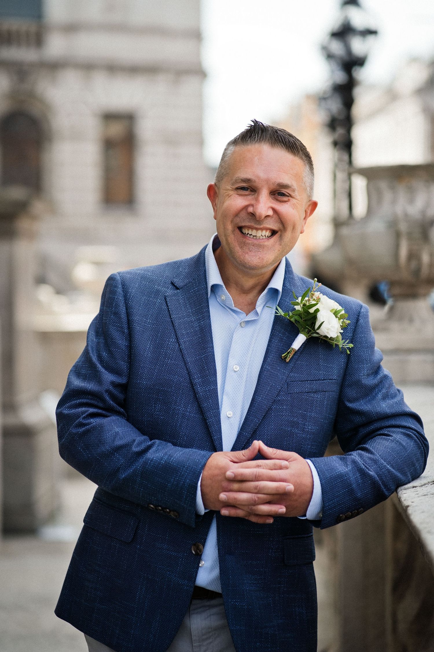 Smiling man in blue blazer with white boutonniere poses outdoors at a wedding ceremony venue.