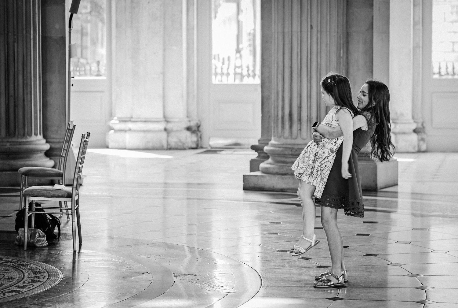 Two young girls stand together on a polished marble floor surrounded by classical columns in a grand historic building.