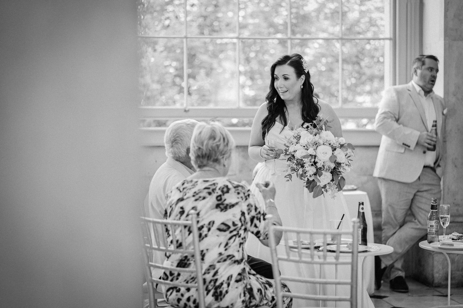 Bride holding bouquet speaks with seated elderly guests at elegant wedding reception in black and white photo.