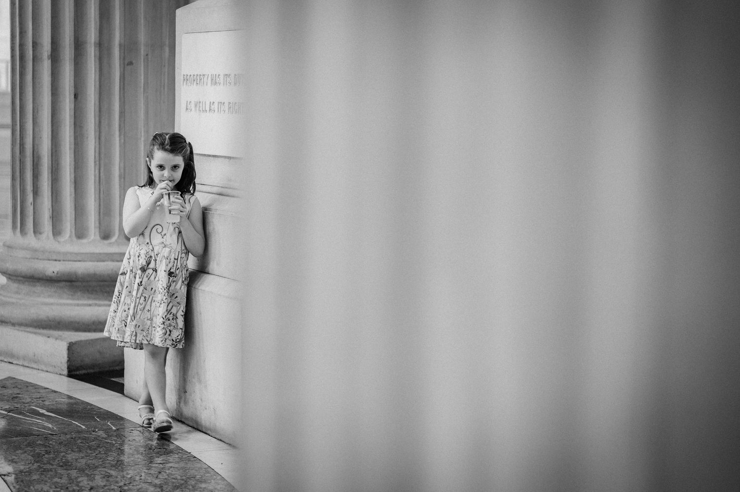 Black and white photo of a young girl in a floral dress leaning against a large stone column.