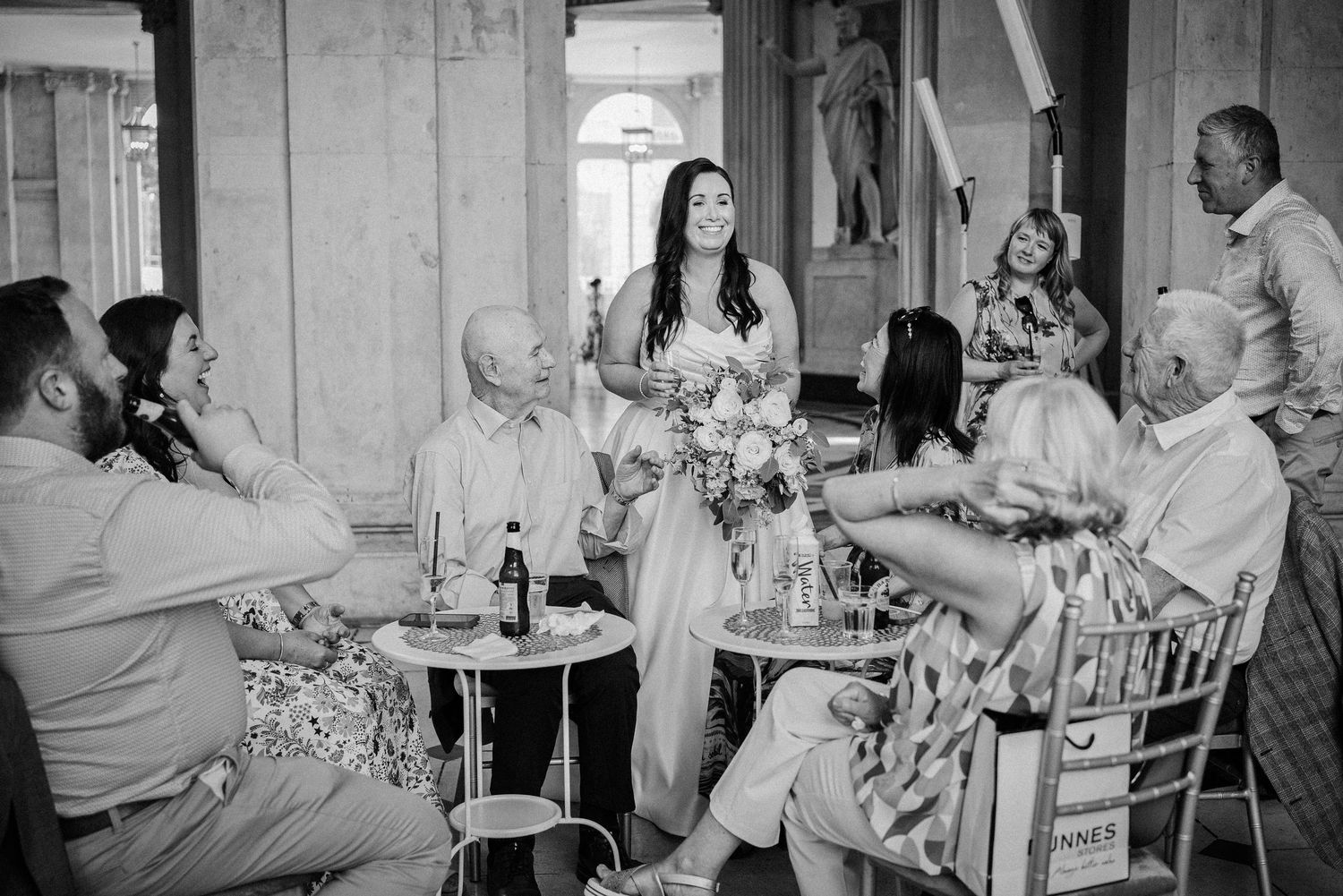 Bride stands holding bouquet addressing seated wedding guests in elegant venue, black and white photo.