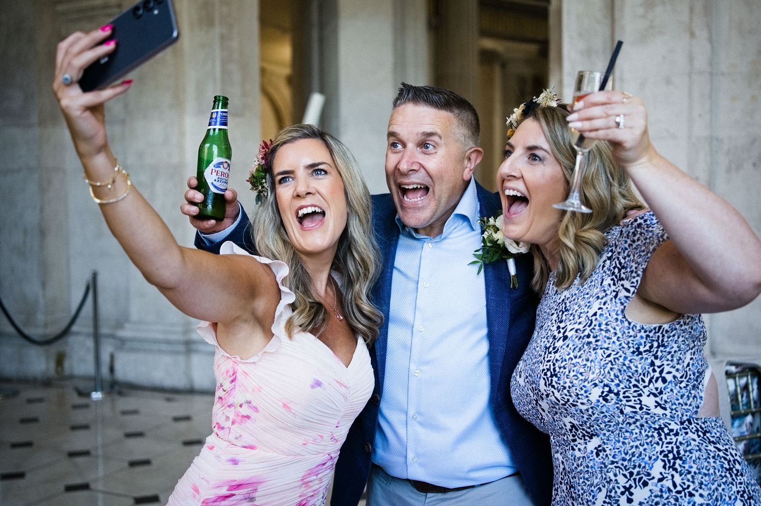 Three wedding guests take a joyful selfie, one holding a green beer bottle, celebrating outdoors.