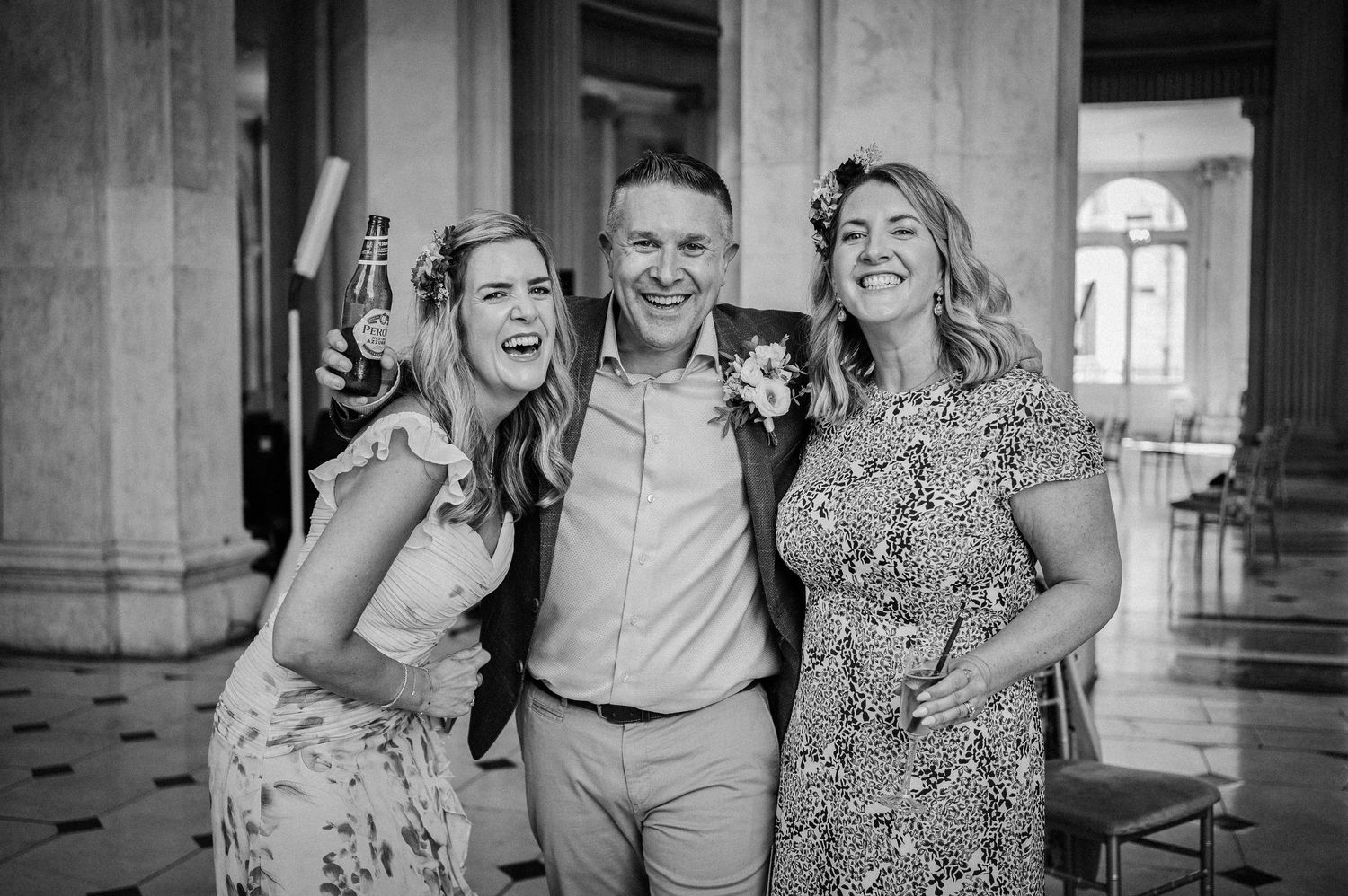 Two women and a man smile together at a wedding celebration in a grand venue, black and white.