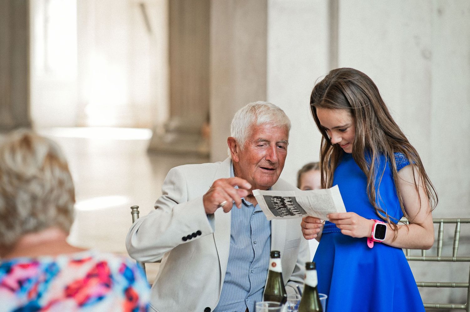 Elderly man and young girl in blue dress reviewing a document together at an elegant indoor event.