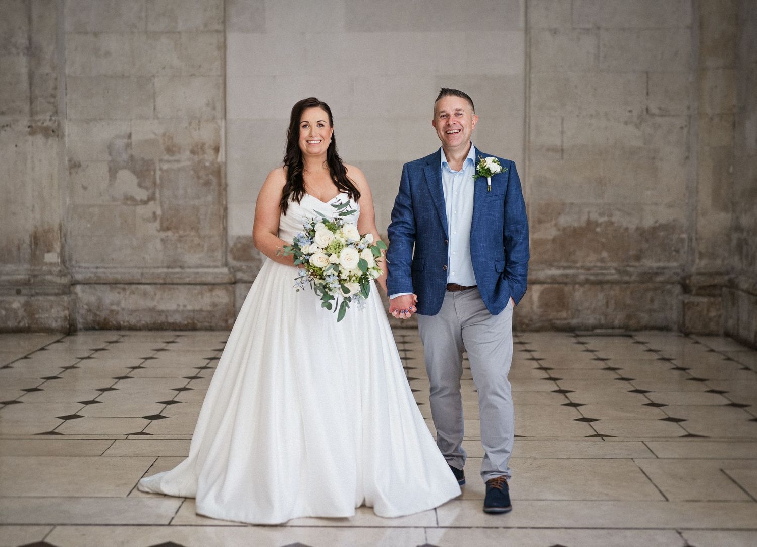 Bride in white gown and groom in blue suit hold hands with a bouquet in an elegant venue.