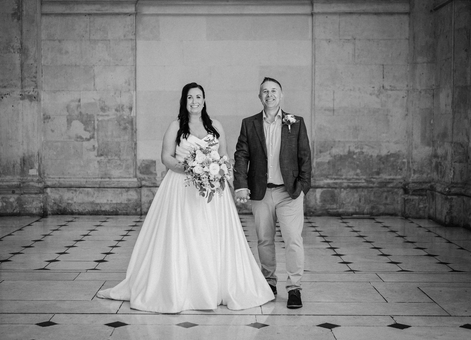 Black and white wedding photo of bride holding bouquet and groom standing together in elegant stone building.