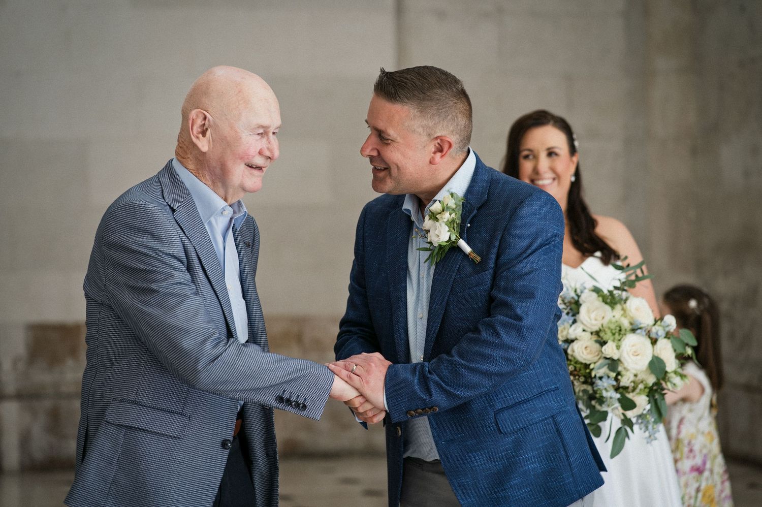 Two men shaking hands and smiling at a wedding, with a bride holding a white bouquet standing behind them.