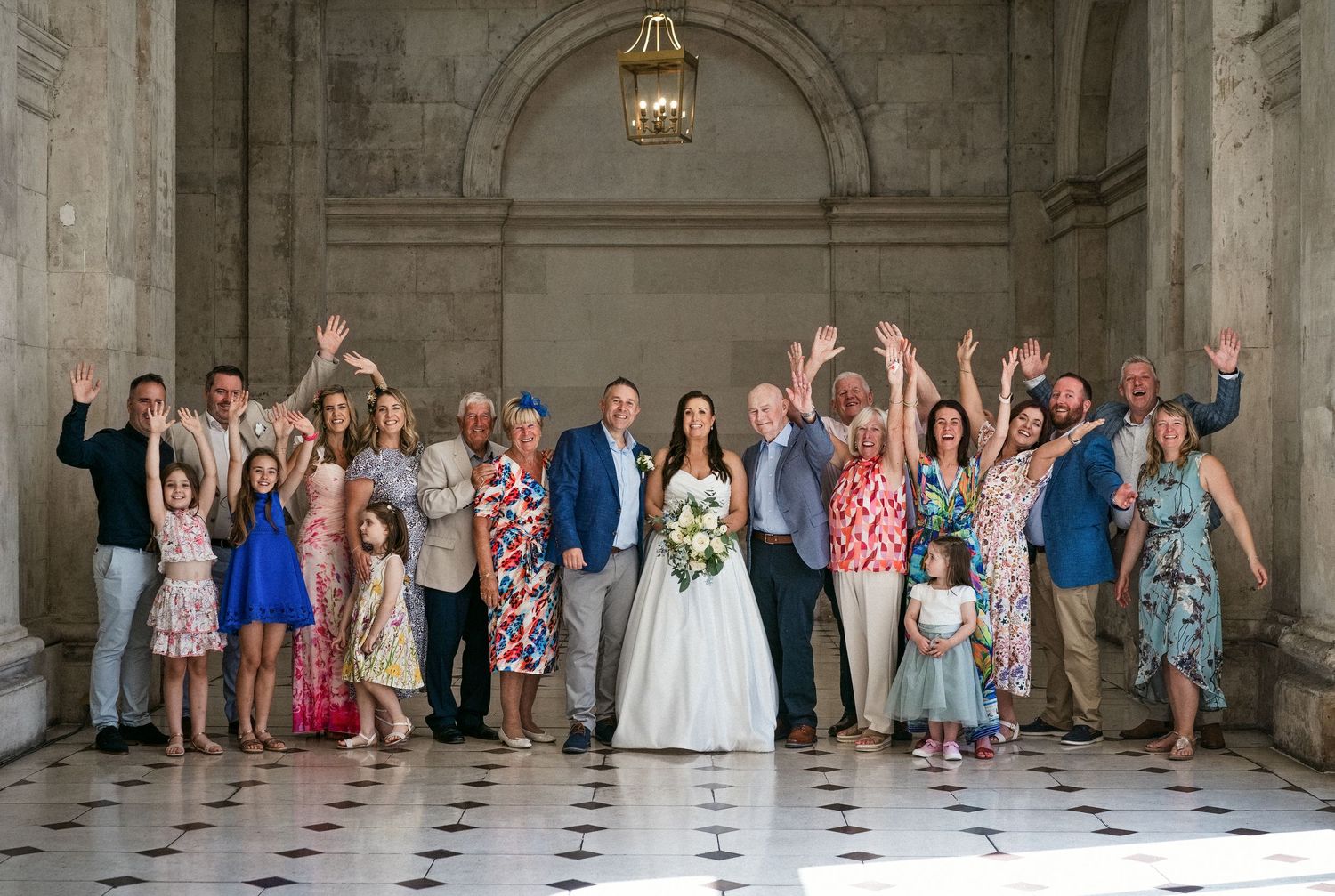 Large wedding group photo with bride in white gown and guests waving hands in elegant marble hall.