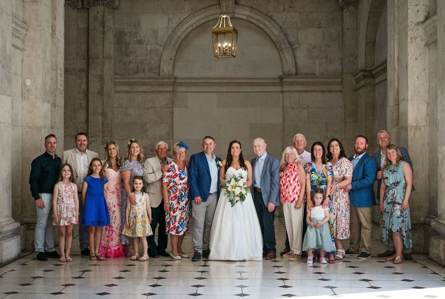Large family group photo at a wedding in an elegant marble hall with ornate architecture.