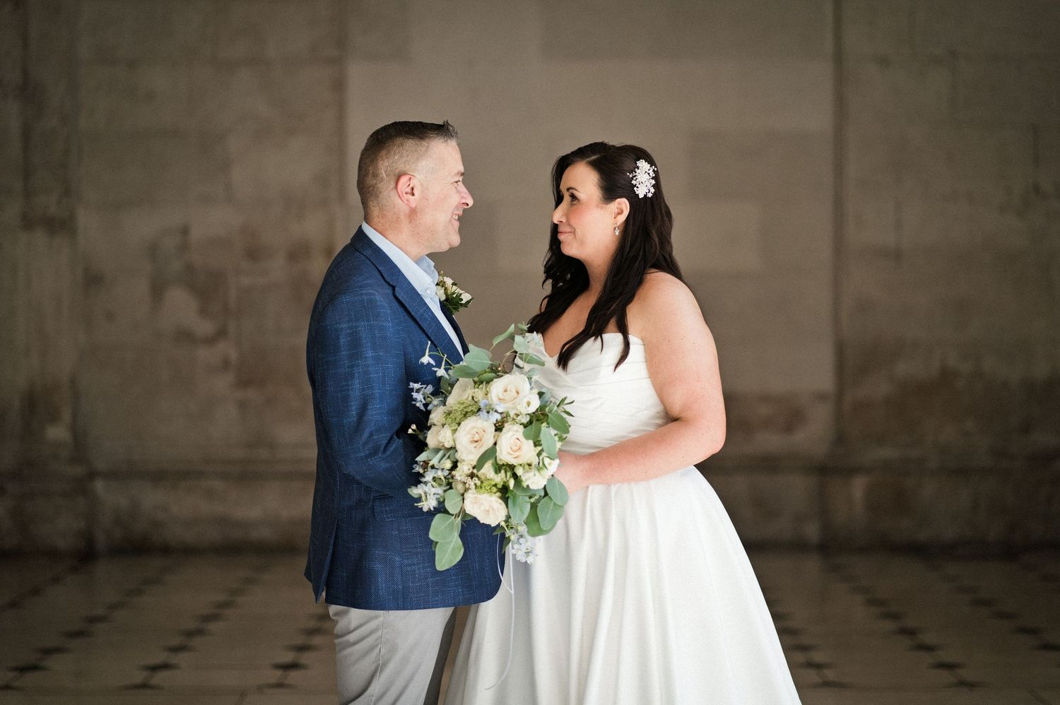 Bride and groom share a romantic moment indoors, bride holding white floral bouquet, groom in blue suit.