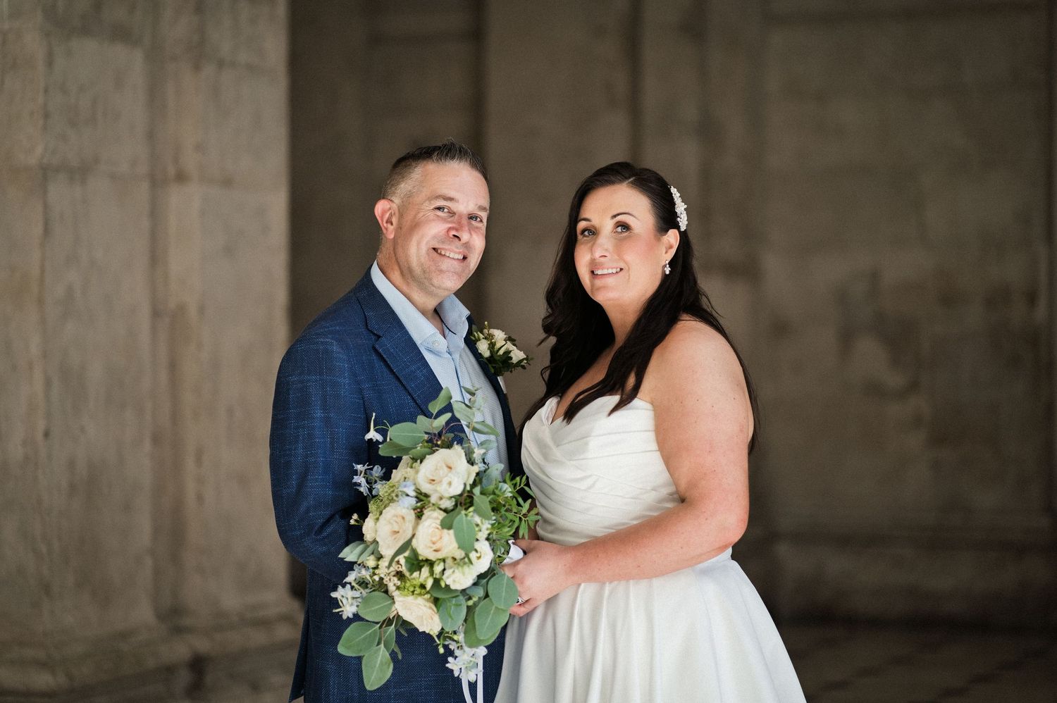 A smiling couple in wedding attire poses with a white floral bouquet against a stone pillar backdrop.