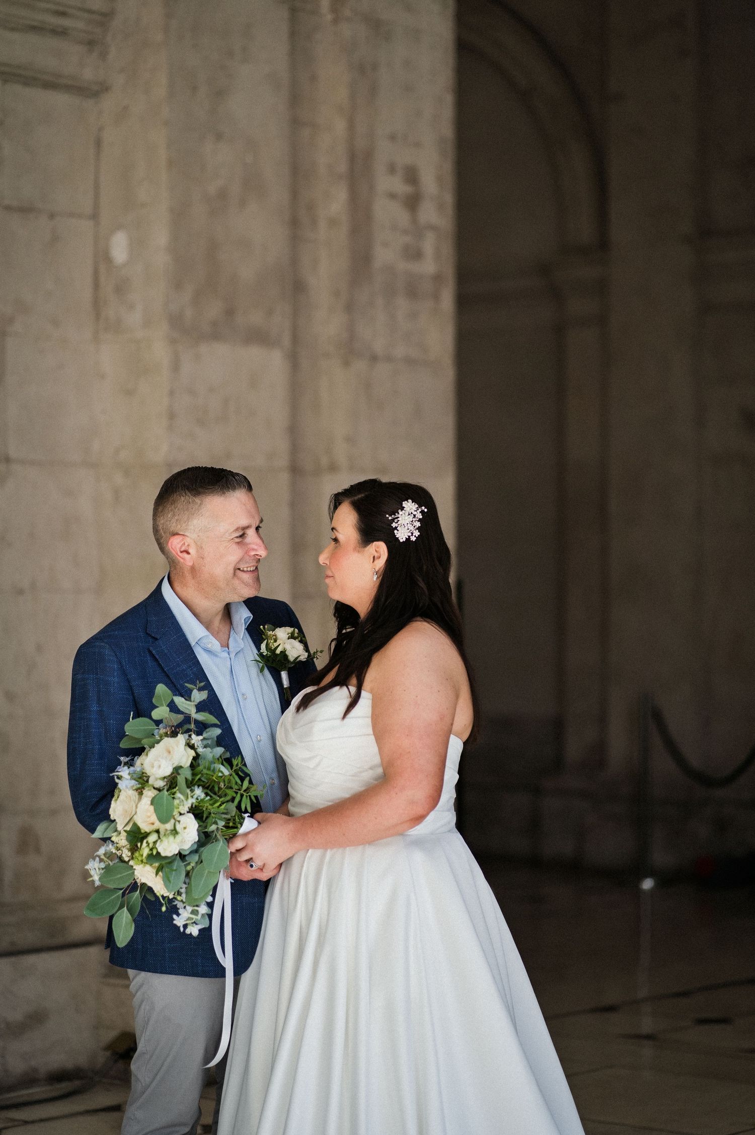 Bride and groom share a romantic moment at a grand stone venue, bride holding white floral bouquet in wedding attire.