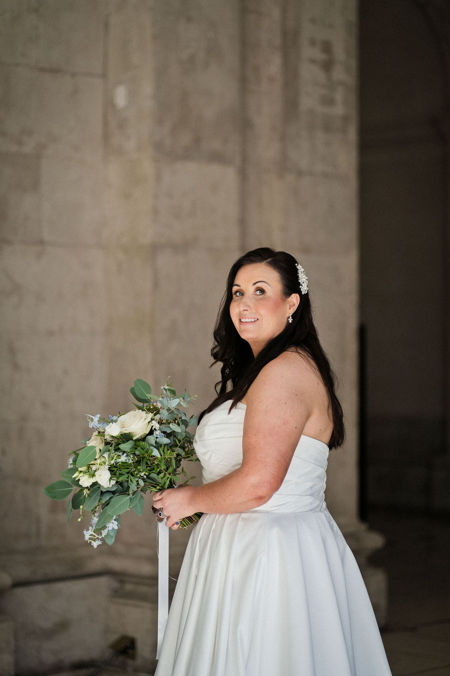 Bride in strapless white gown holding green and white bouquet, smiling beside stone columns.