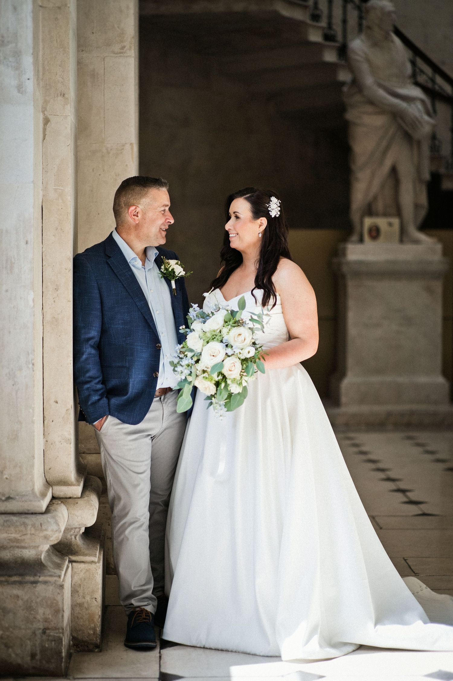 Bride in white gown holding bouquet and groom in navy blazer share a loving glance at an elegant wedding venue.