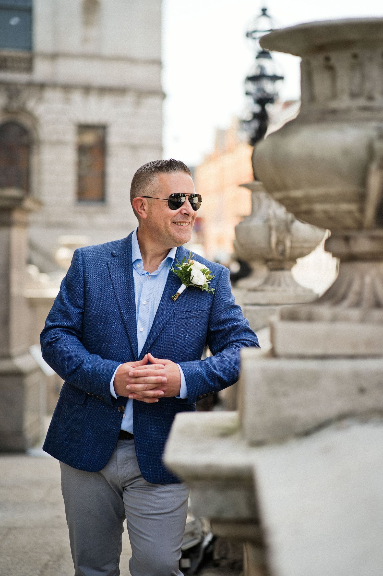 Smiling groom in blue blazer with boutonniere and aviator sunglasses posing near stone fountain outdoors.