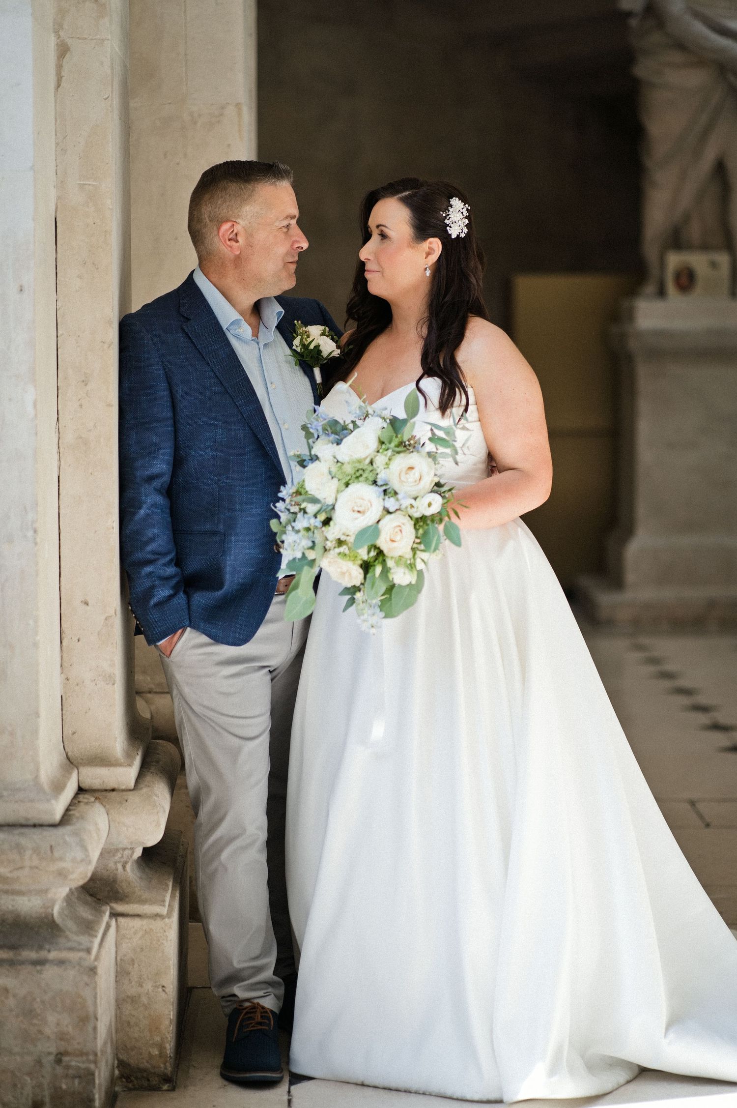 A bride in a white gown holding a white bouquet stands with a groom in a navy blazer among stone columns.