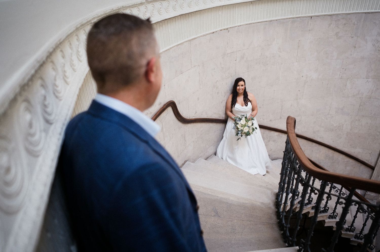 Bride in white gown holds bouquet on grand staircase as groom in blue suit looks up at her during wedding.