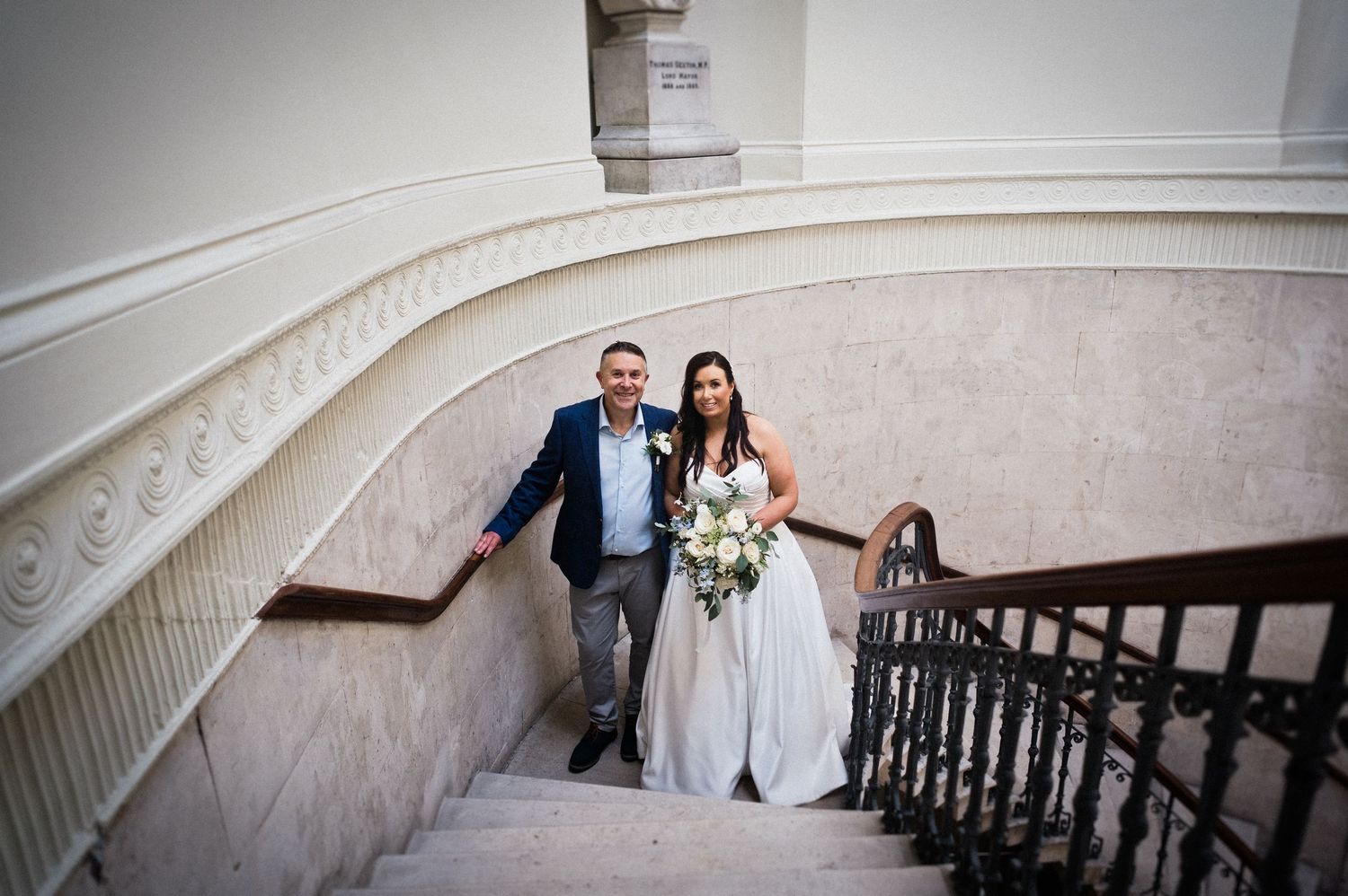 Bride and groom walking down elegant marble staircase with ornate iron railing, bride holding white bouquet.