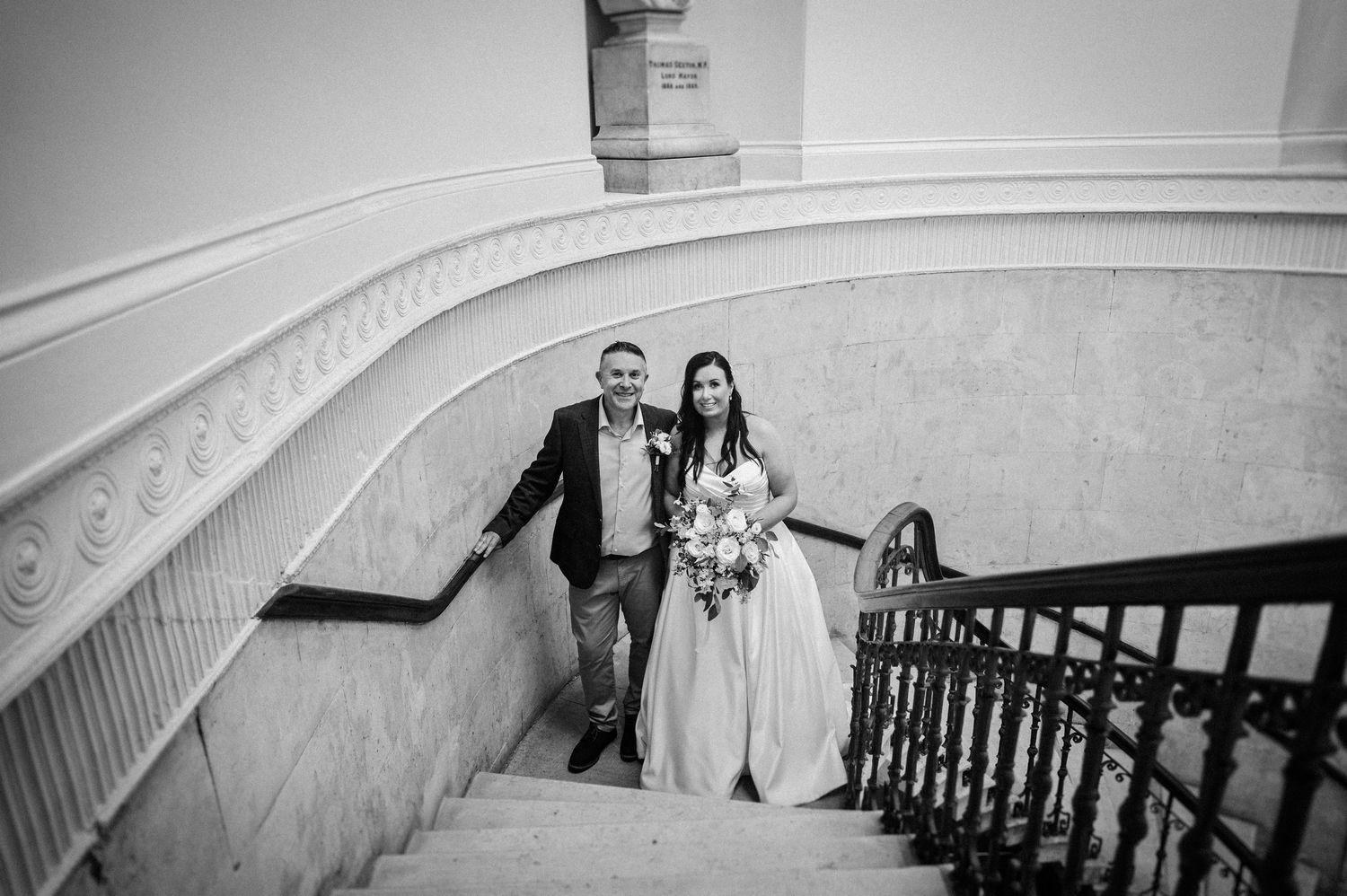 Black and white photo of a bride and groom walking down an elegant curved staircase on their wedding day.