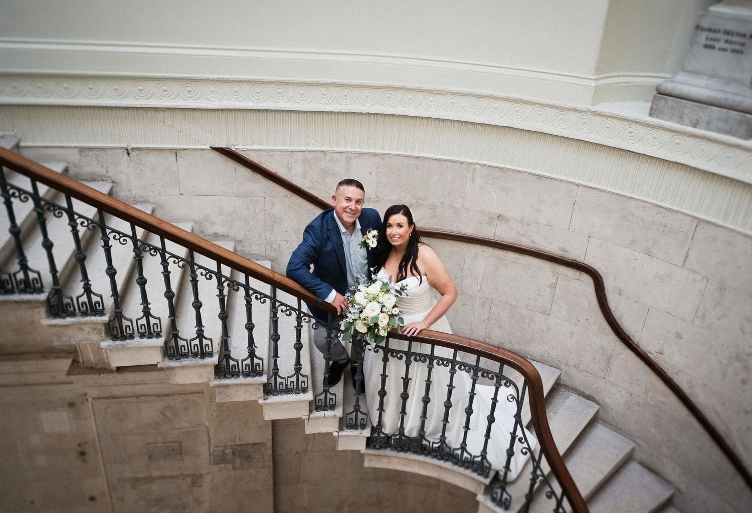 Bride and groom posing on a grand curved staircase with ornate iron railings, bride holding white bouquet.