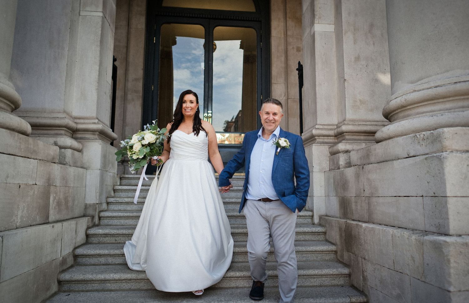 Bride and groom walking down outdoor stone steps between classical columns after their wedding ceremony.