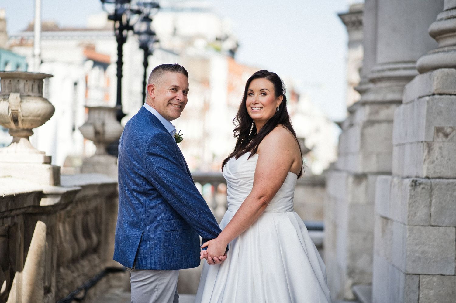 Bride and groom hold hands smiling on a historic stone balcony, groom in blue suit, bride in white strapless gown.