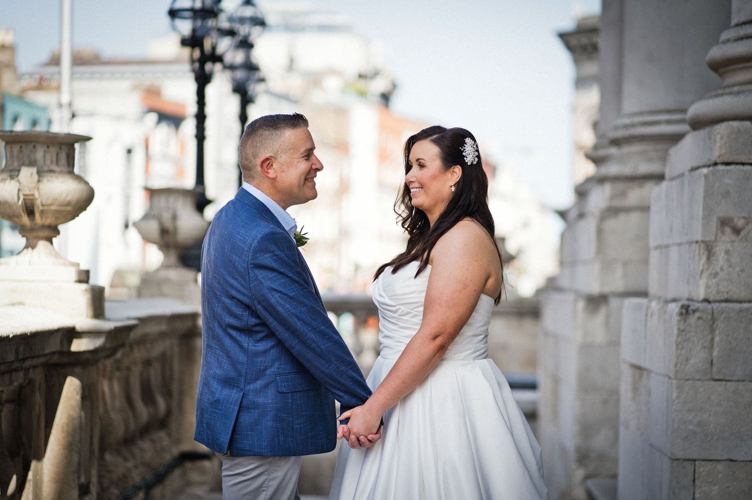 A bride and groom hold hands and gaze at each other during outdoor wedding photos near classic stone columns.
