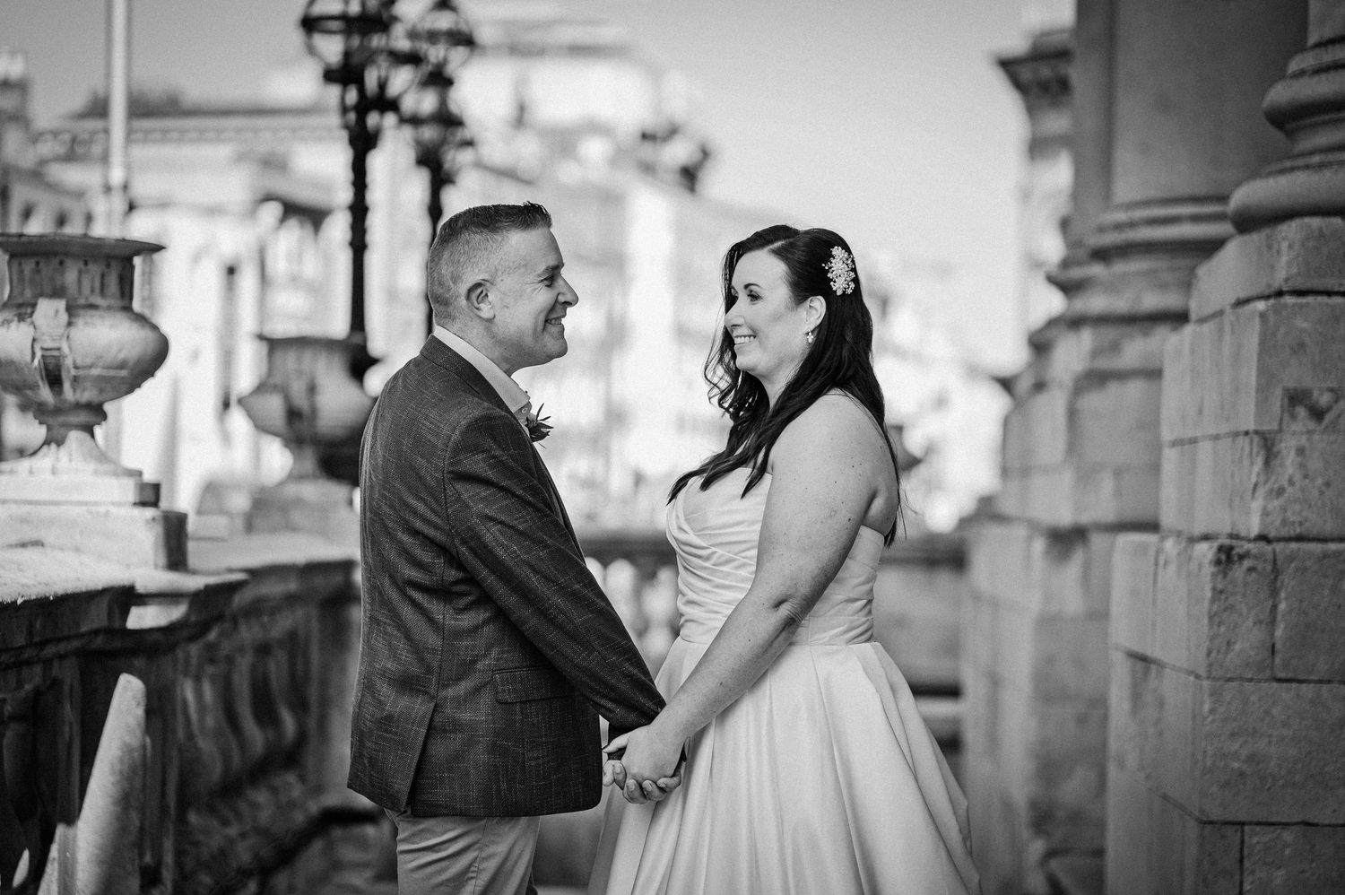 Black and white wedding photo of a couple holding hands and gazing at each other near elegant stone columns.