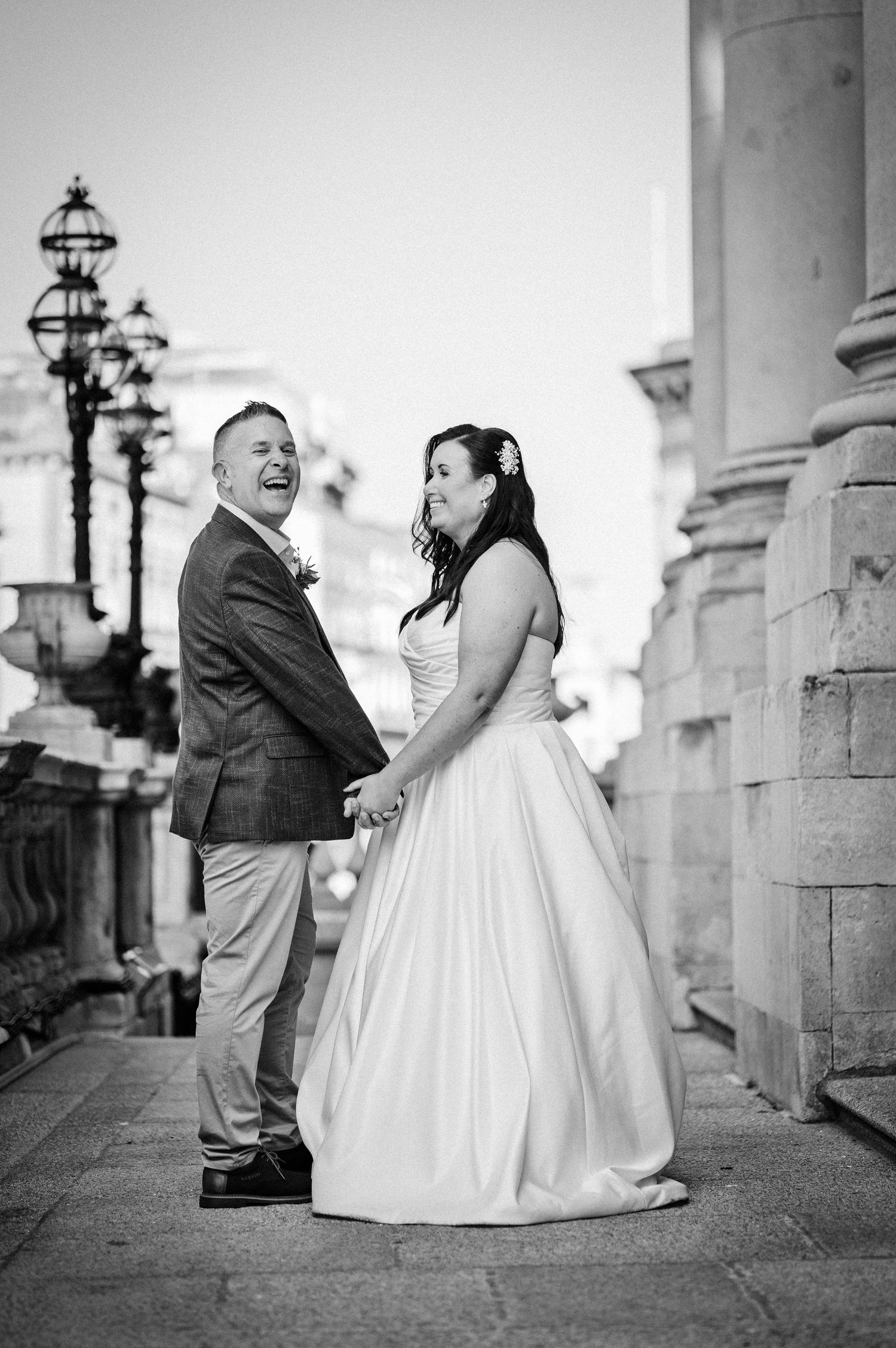 Black and white photo of a laughing bride and groom holding hands on a cobblestone street during their wedding day.