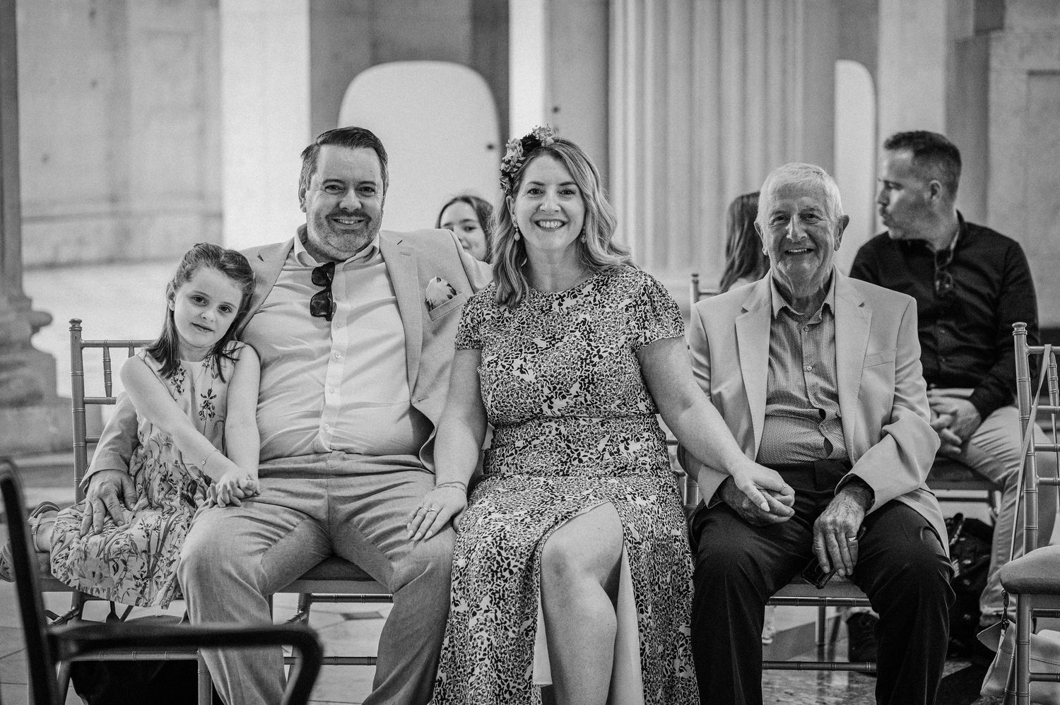 Black and white photo of a smiling family seated together at an elegant outdoor ceremony with columns.
