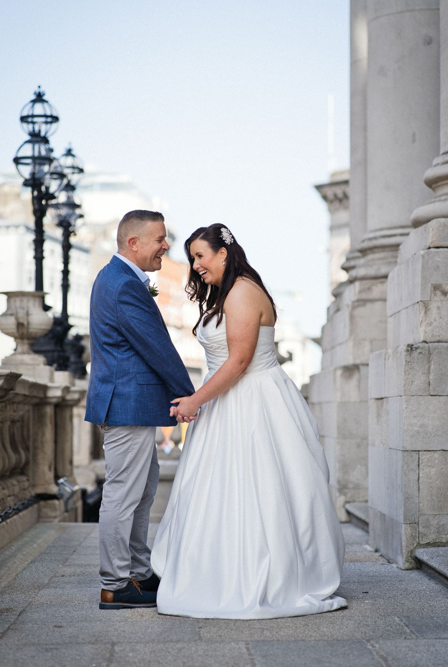 Bride and groom holding hands and smiling at each other in front of elegant stone columns on their wedding day.
