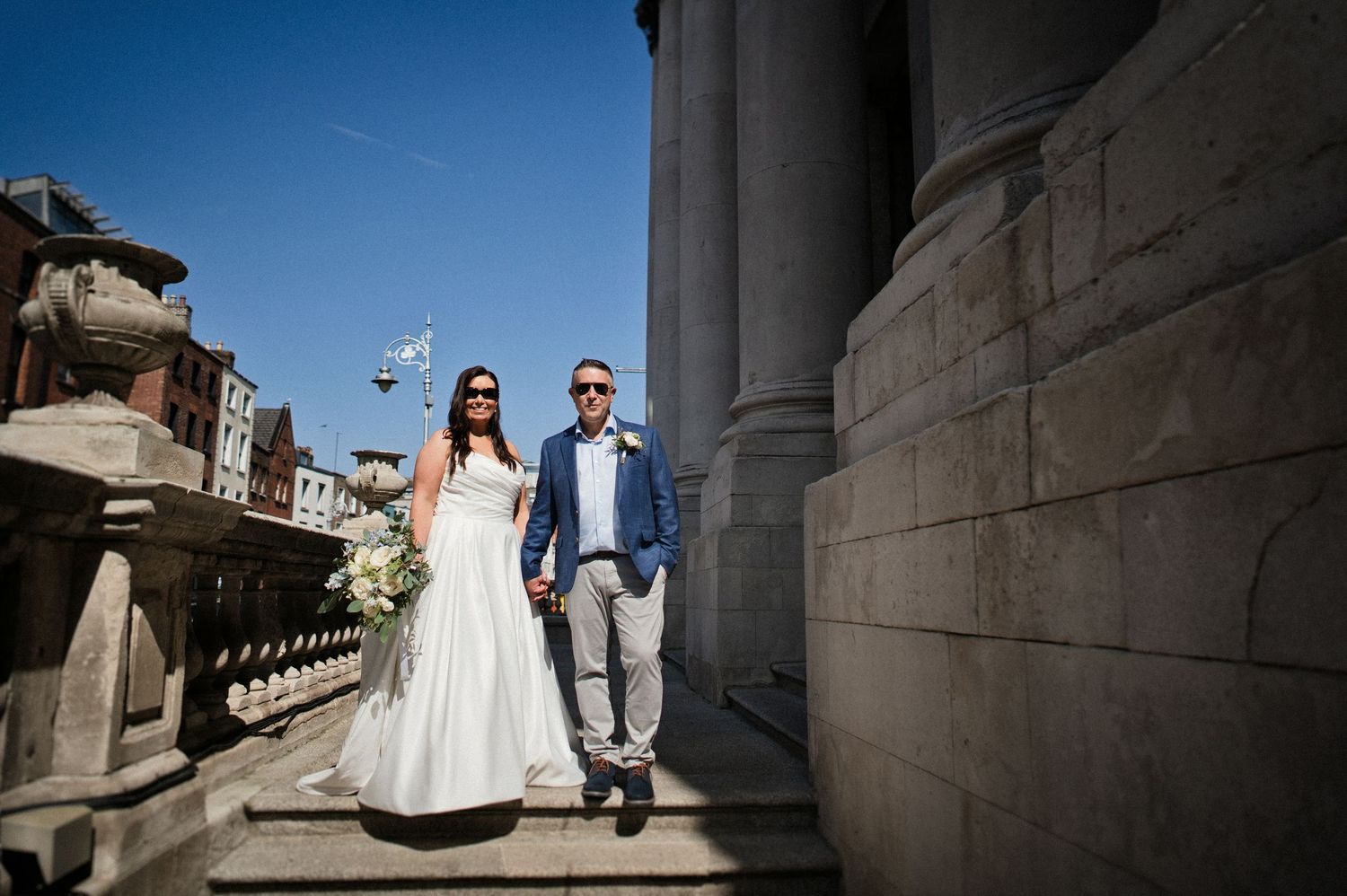 Bride and groom pose on stone steps of a grand building under a clear blue sky on their wedding day.