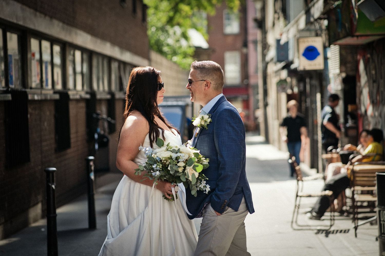 Bride and groom standing together on a sunny urban street, bride holding white bouquet, groom in navy blazer.