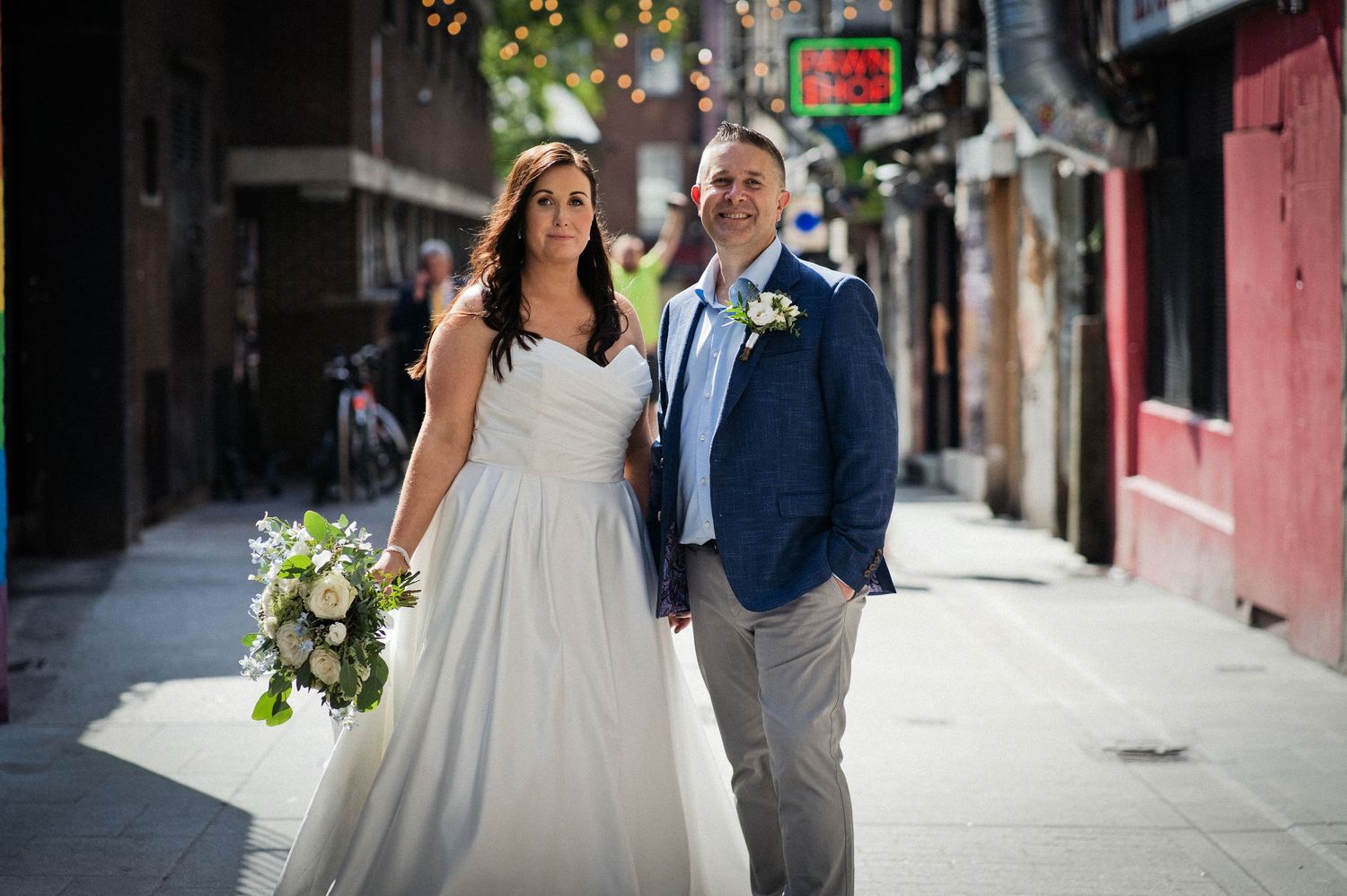 Bride and groom walking hand in hand down a sunny urban alley, bride holding a white bouquet in a strapless gown.
