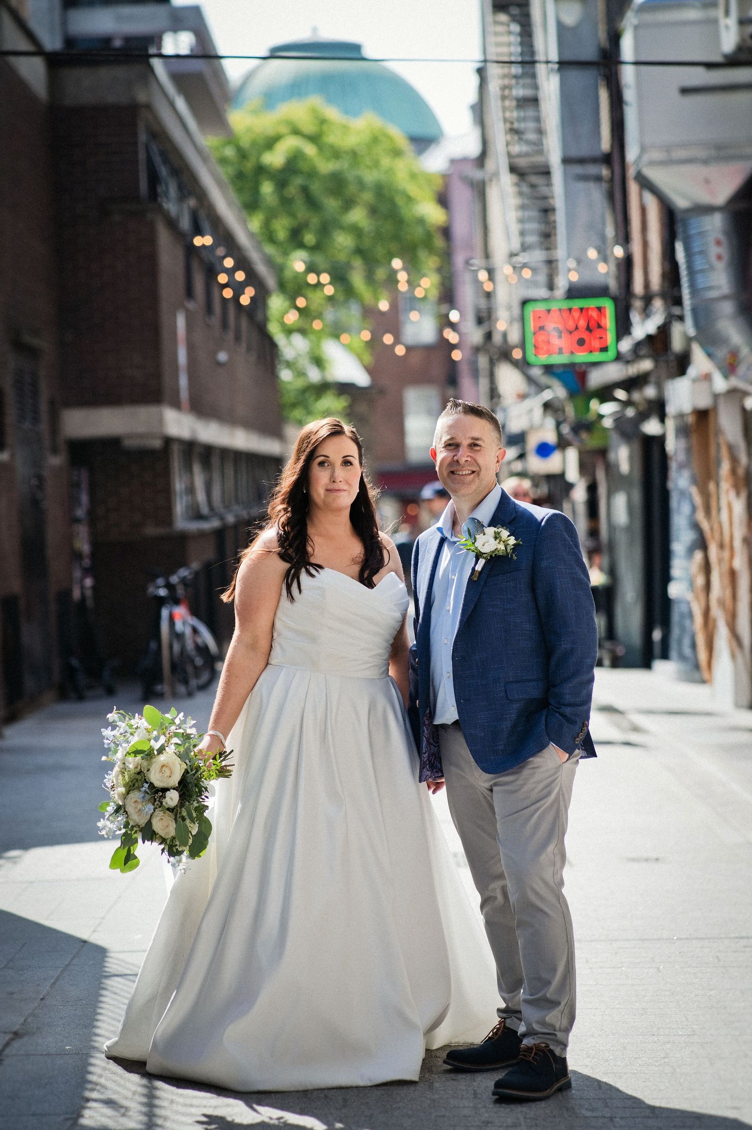 Bride in white gown holding bouquet stands with groom in blue suit on a sunny urban street with string lights.