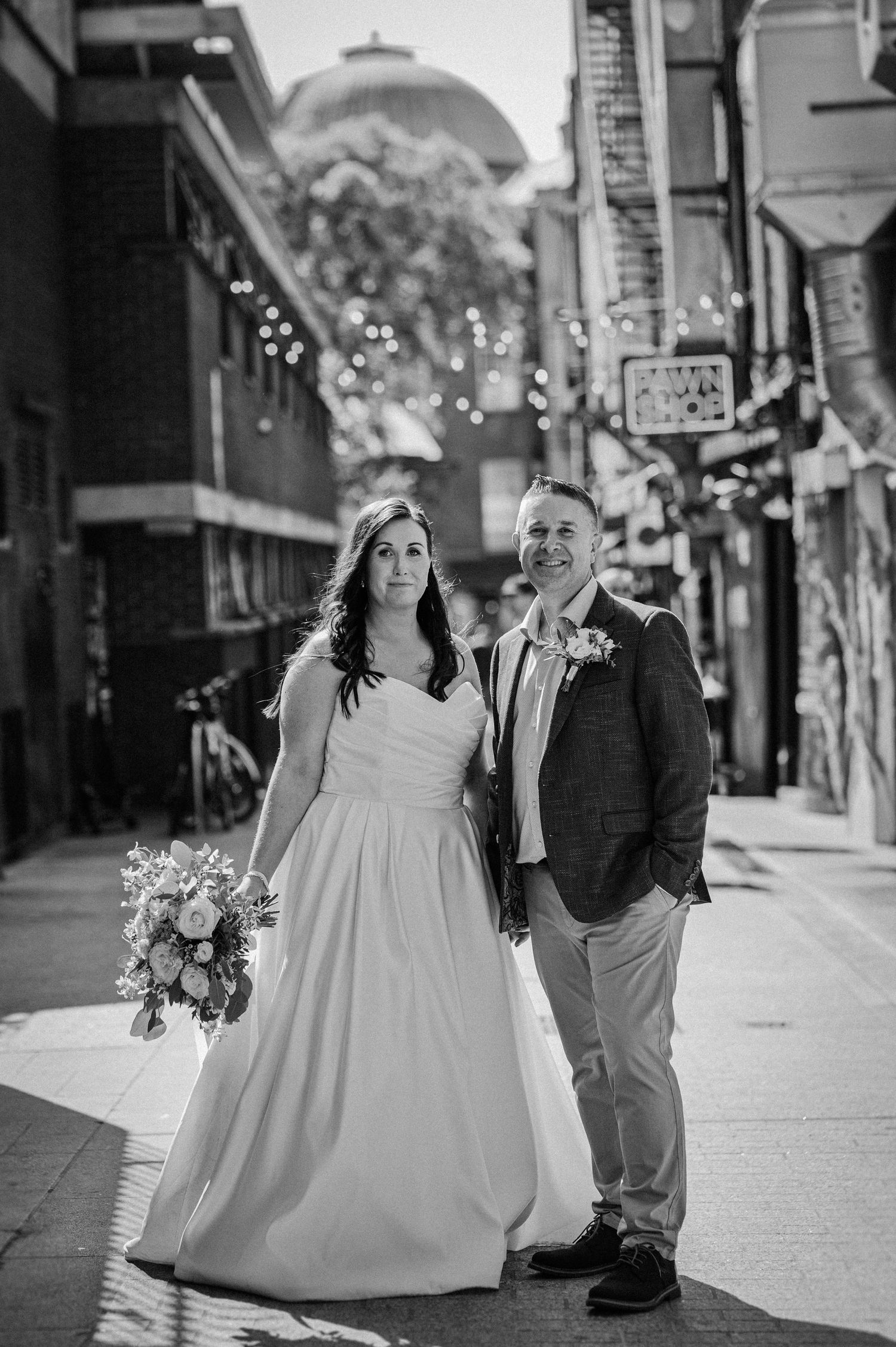 Black and white photo of a bride in white gown and groom in suit posing on a city sidewalk with string lights.