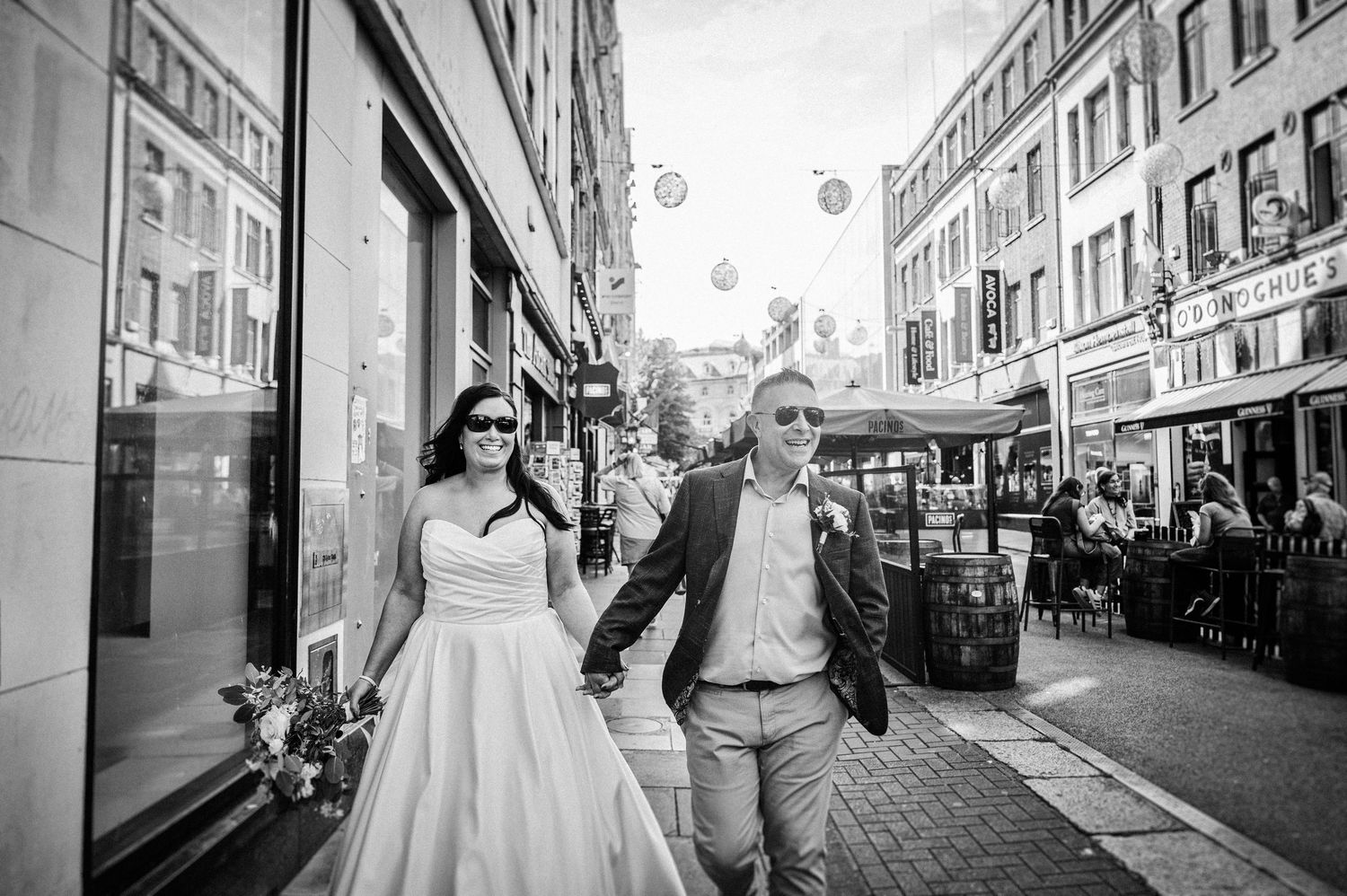 Bride and groom walk hand-in-hand down a bustling city street in black and white wedding photo.