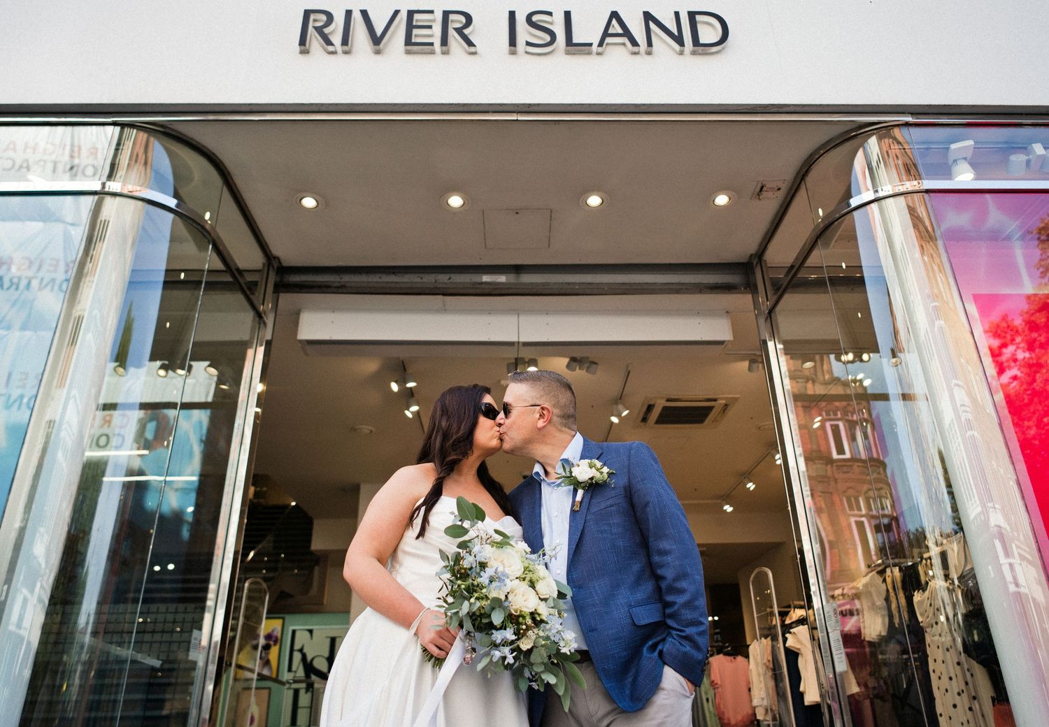 Bride and groom kissing outside a River Island store, bride holding a white floral bouquet in a wedding dress.
