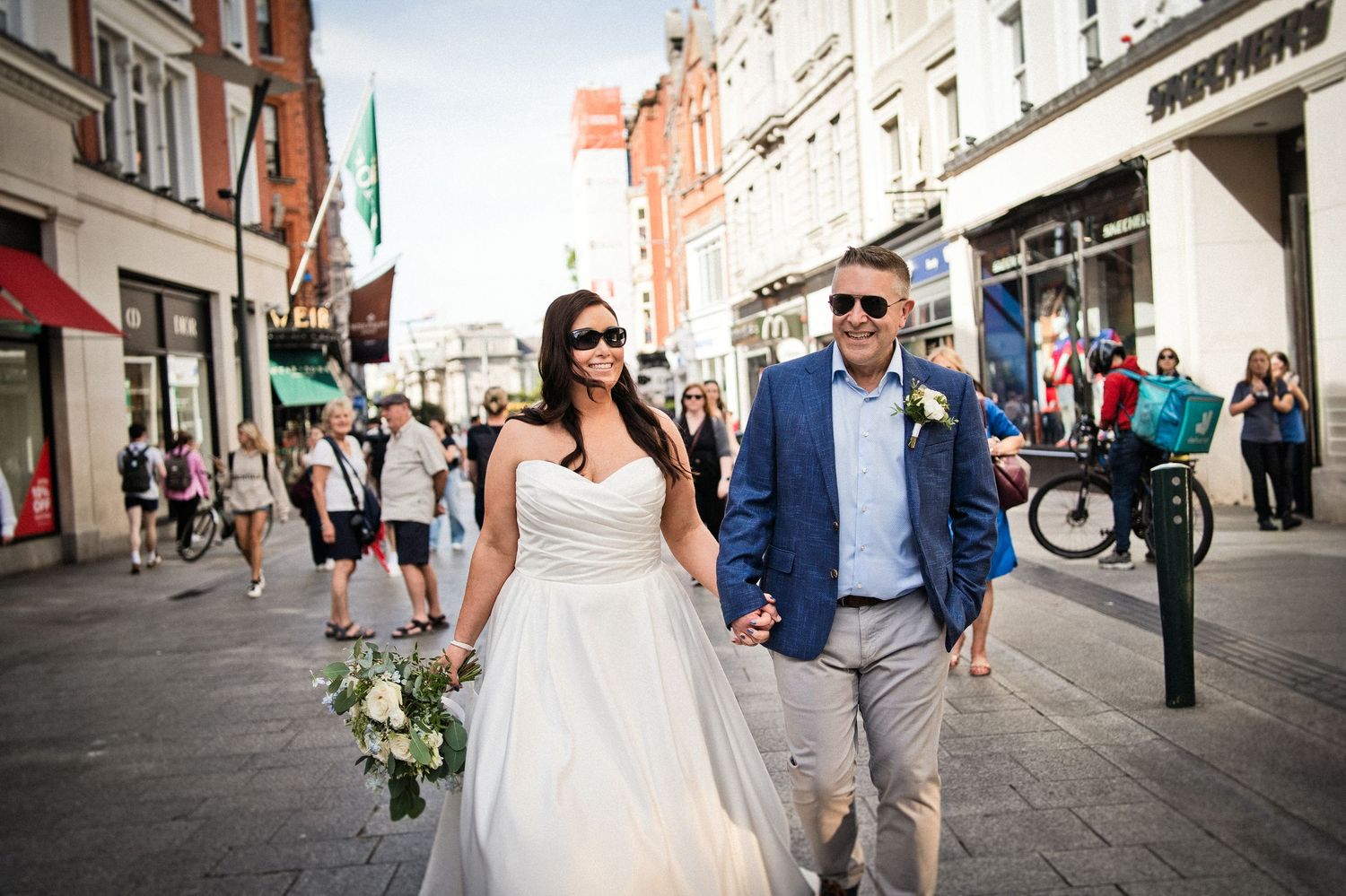Bride and groom walking hand-in-hand on a sunny city street, bride holding a bouquet, both wearing sunglasses.