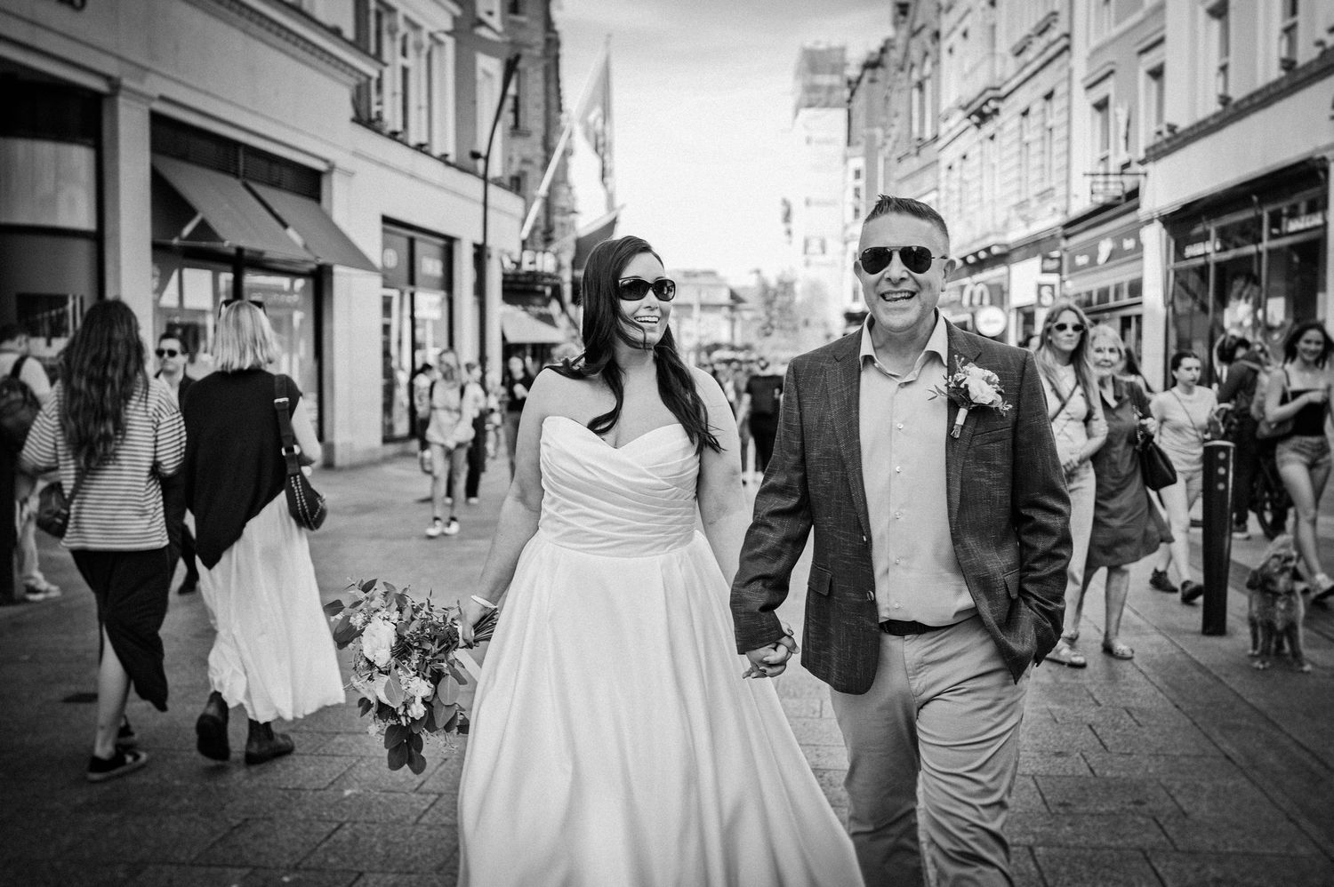 Bride and groom walking hand in hand through a busy street, smiling and wearing sunglasses, in black and white.